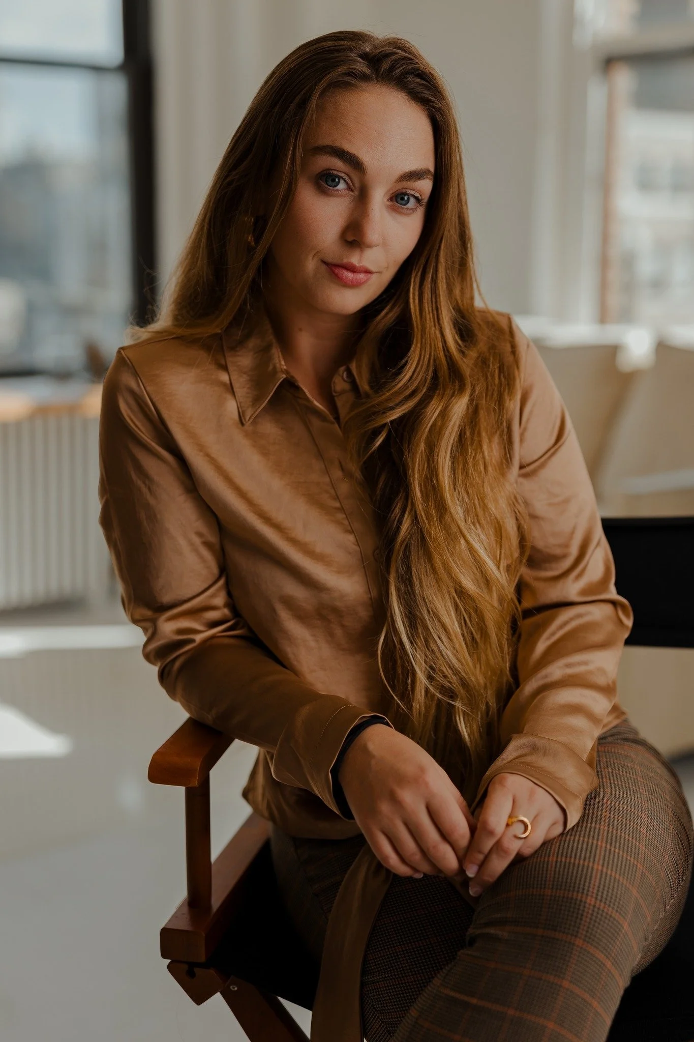 Portrait of a young woman with long red hair, blue eyes, sitting indoors in a cozy, well-lit room with large windows.