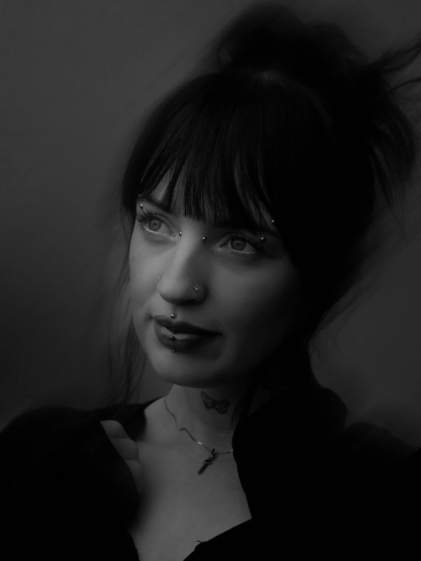 Black and white portrait of a woman with multiple facial piercings, dark makeup, and tattoos, with dark hair and bangs, looking slightly upwards and to the side.