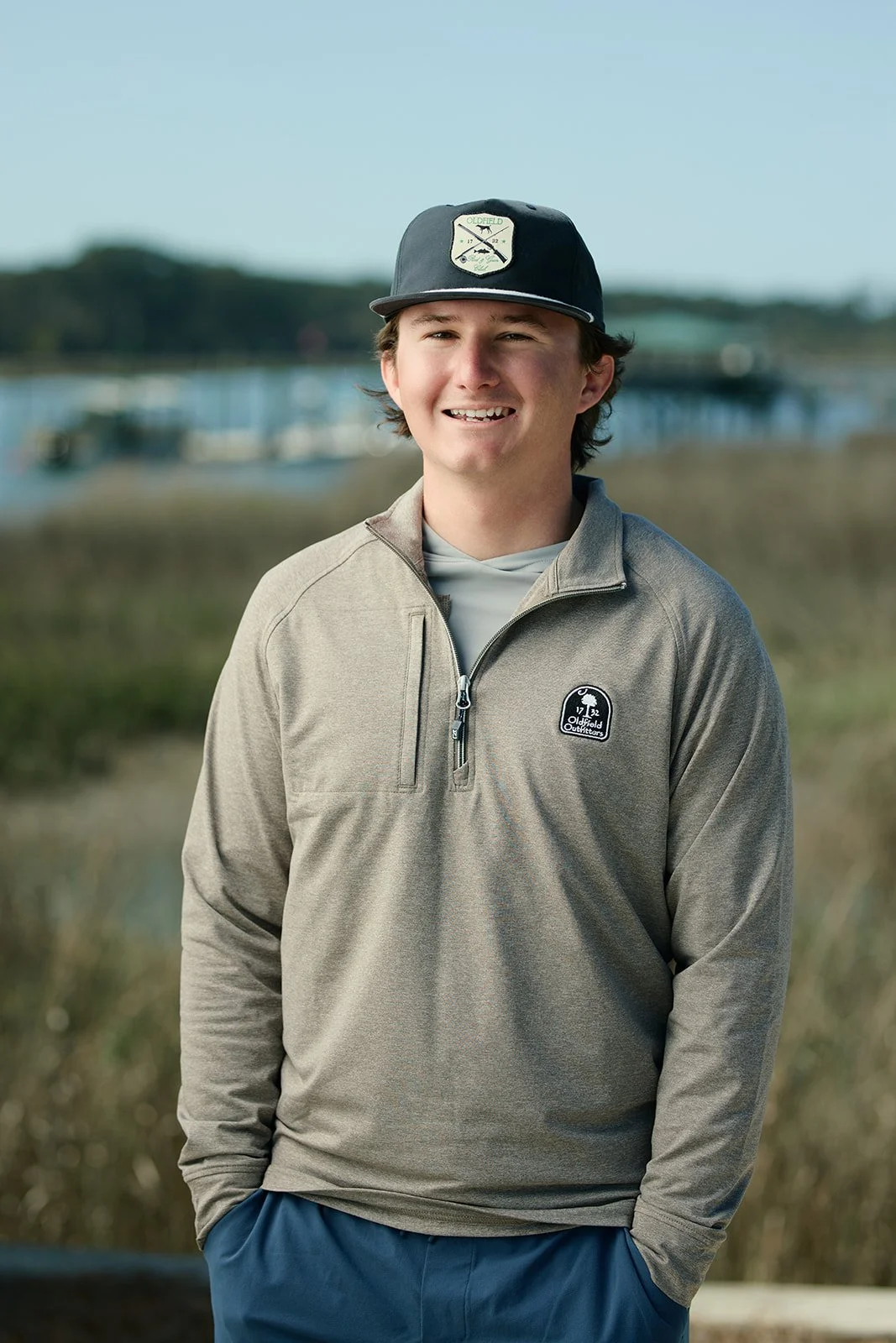 A young man wearing a gray quarter-zip jacket and a black hat with a logo stands outdoors near water and grass, smiling at the camera.