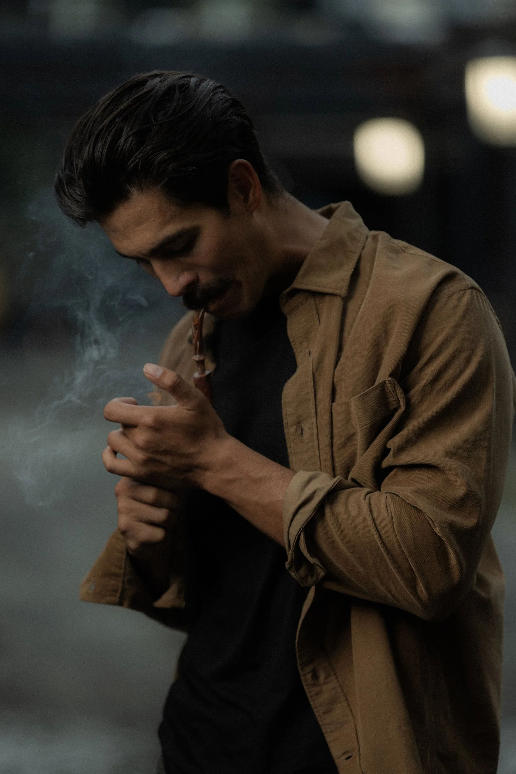 A man with dark hair and a mustache, wearing a tan jacket over a black shirt, is smoking a pipe with smoke coming out, looking down thoughtfully in a dark, softly lit environment.