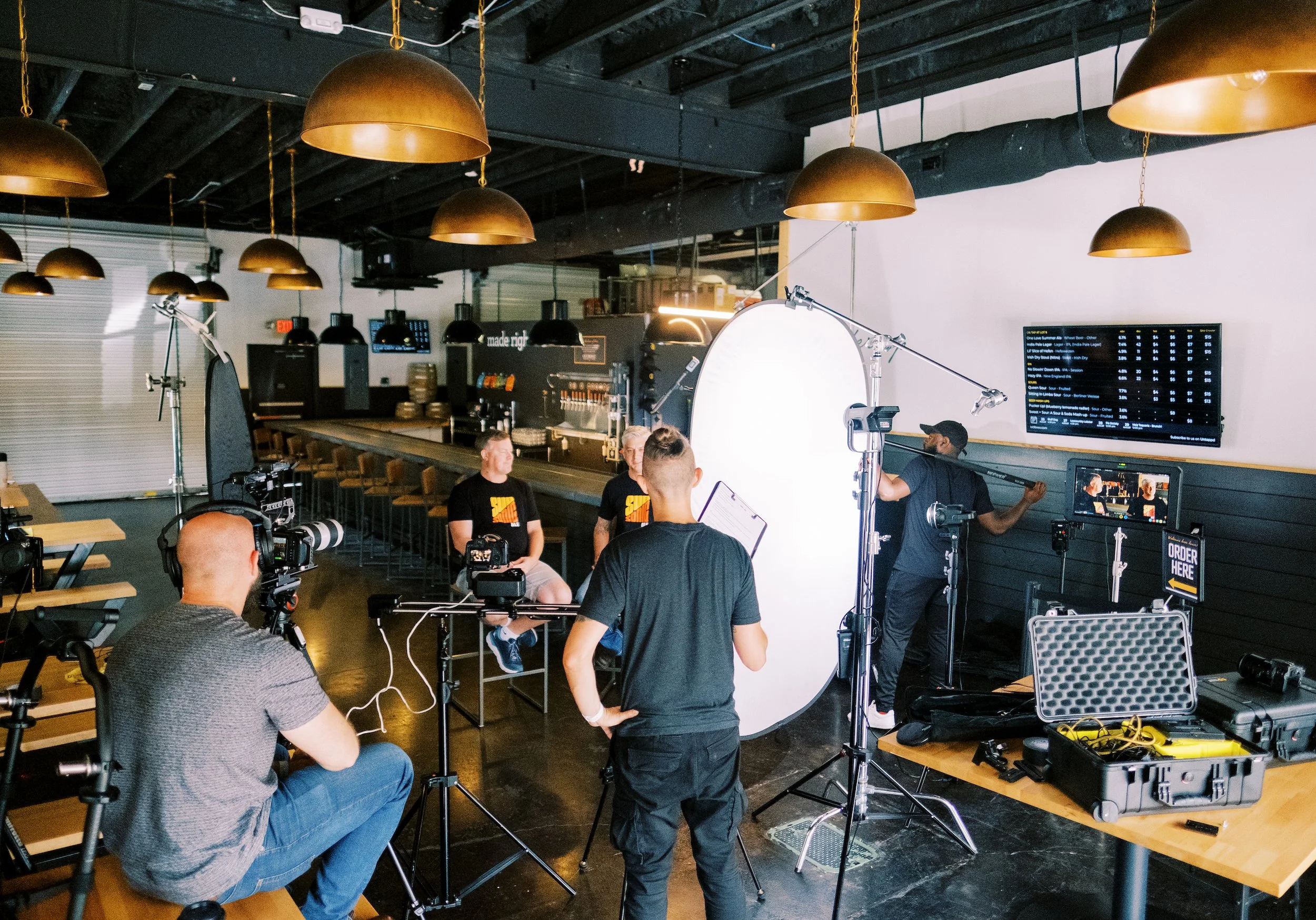 A video shoot inside a modern restaurant or bar with crew members setting up cameras and lighting equipment, with two individuals sitting for the shoot against a bar counter and multiple pendant lights hanging from a black ceiling.