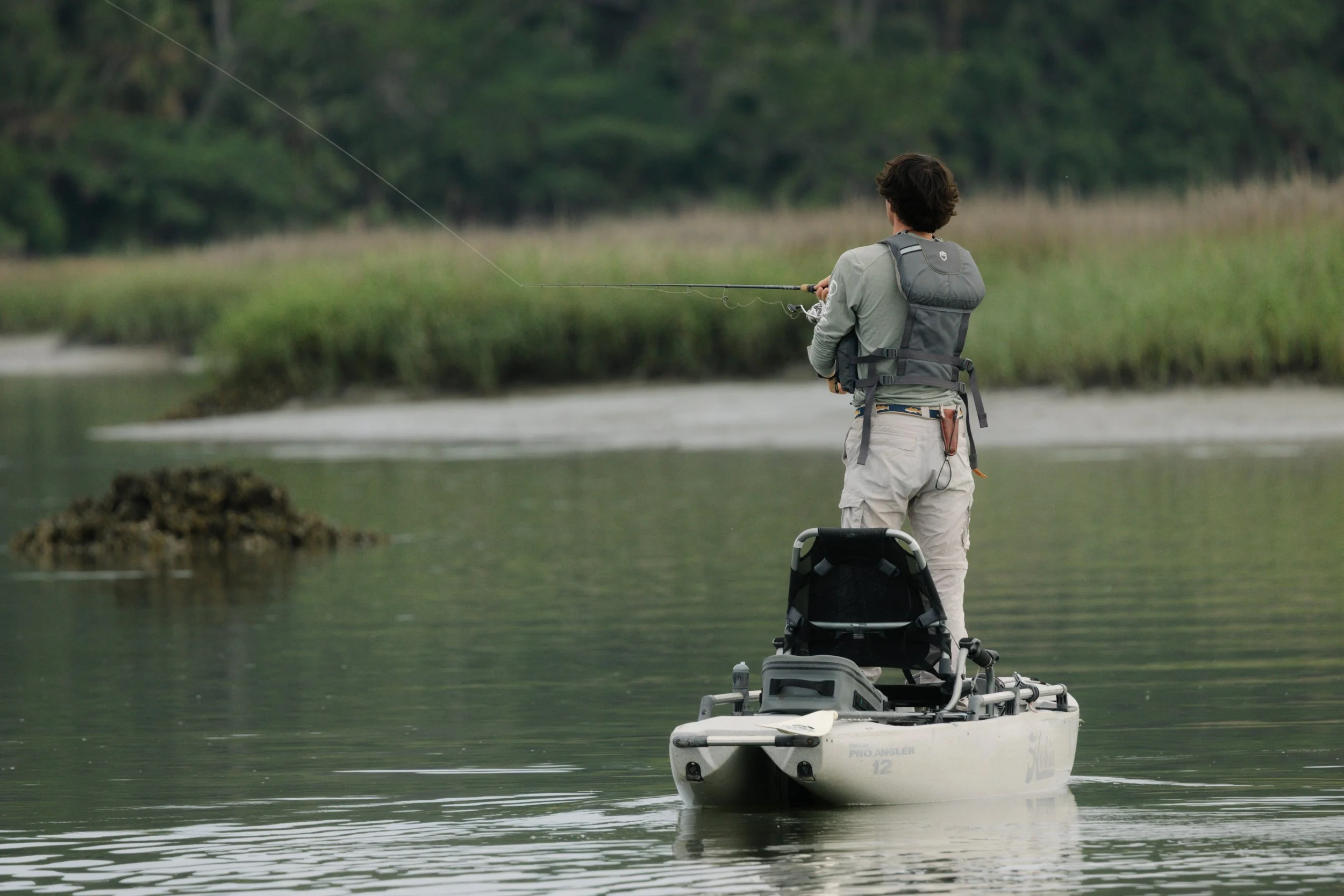 Person fishing from a kayak on a calm river in a natural setting with greenery and trees in the background.