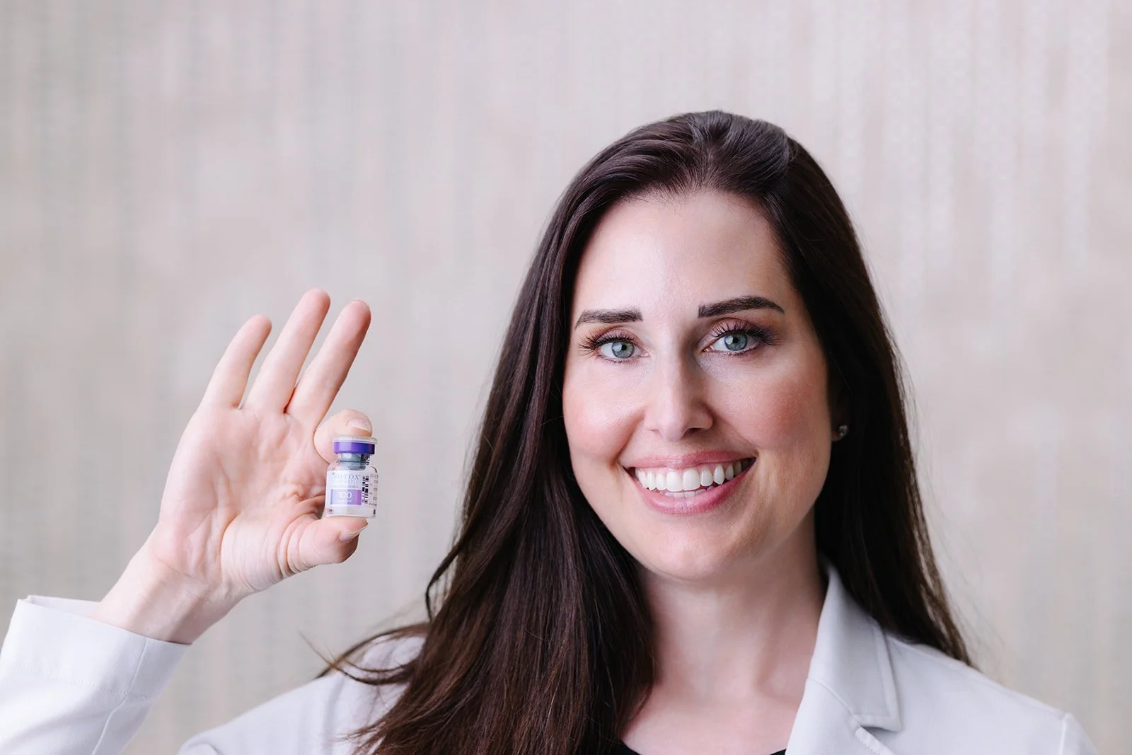 A woman with long dark hair smiling and holding a small vaccine vial in her right hand, displaying it towards the camera against a plain background.