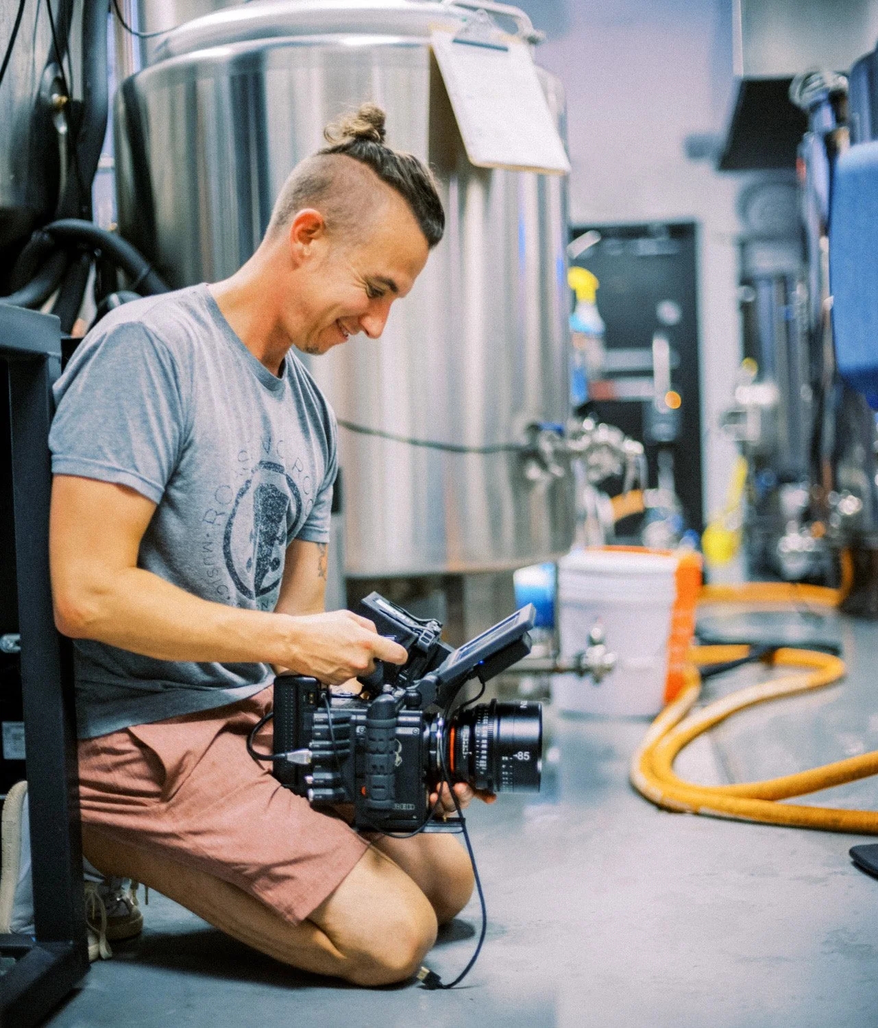 Man kneeling on the floor in a brewery, holding a professional video camera, surrounded by brewery equipment and hoses, smiling and looking at the camera.