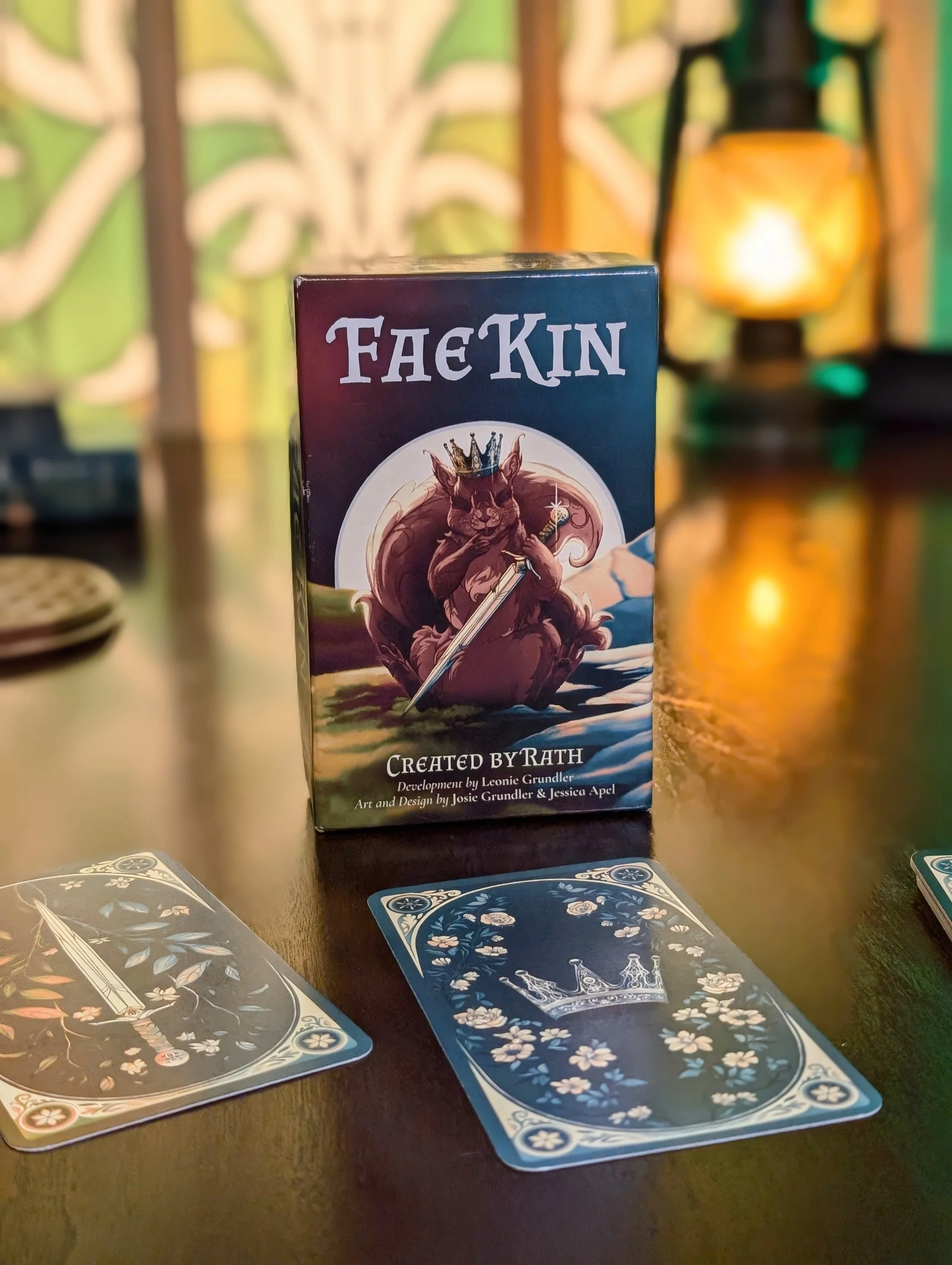 A card game named 'Fae Kin' with a box featuring a squirrel with a crown holding a dagger.. Two face-up cards are visible on the table, one with a dagger and the other with a crown featuring elaborate floral designs.