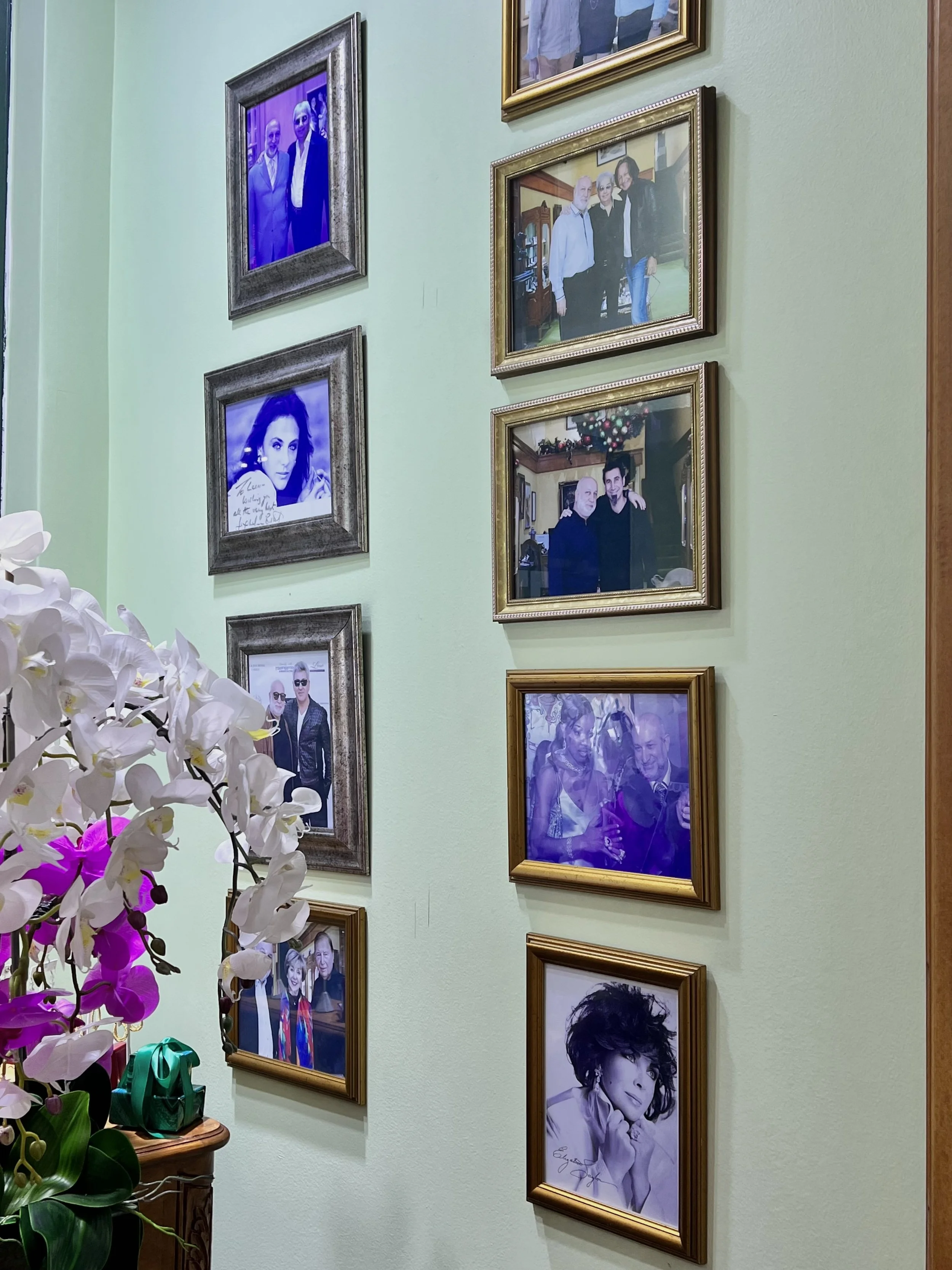 A wall decorated with multiple framed photographs of people, some in color and some in black and white, in a home or gallery setting. There are also white and purple orchids in the foreground.