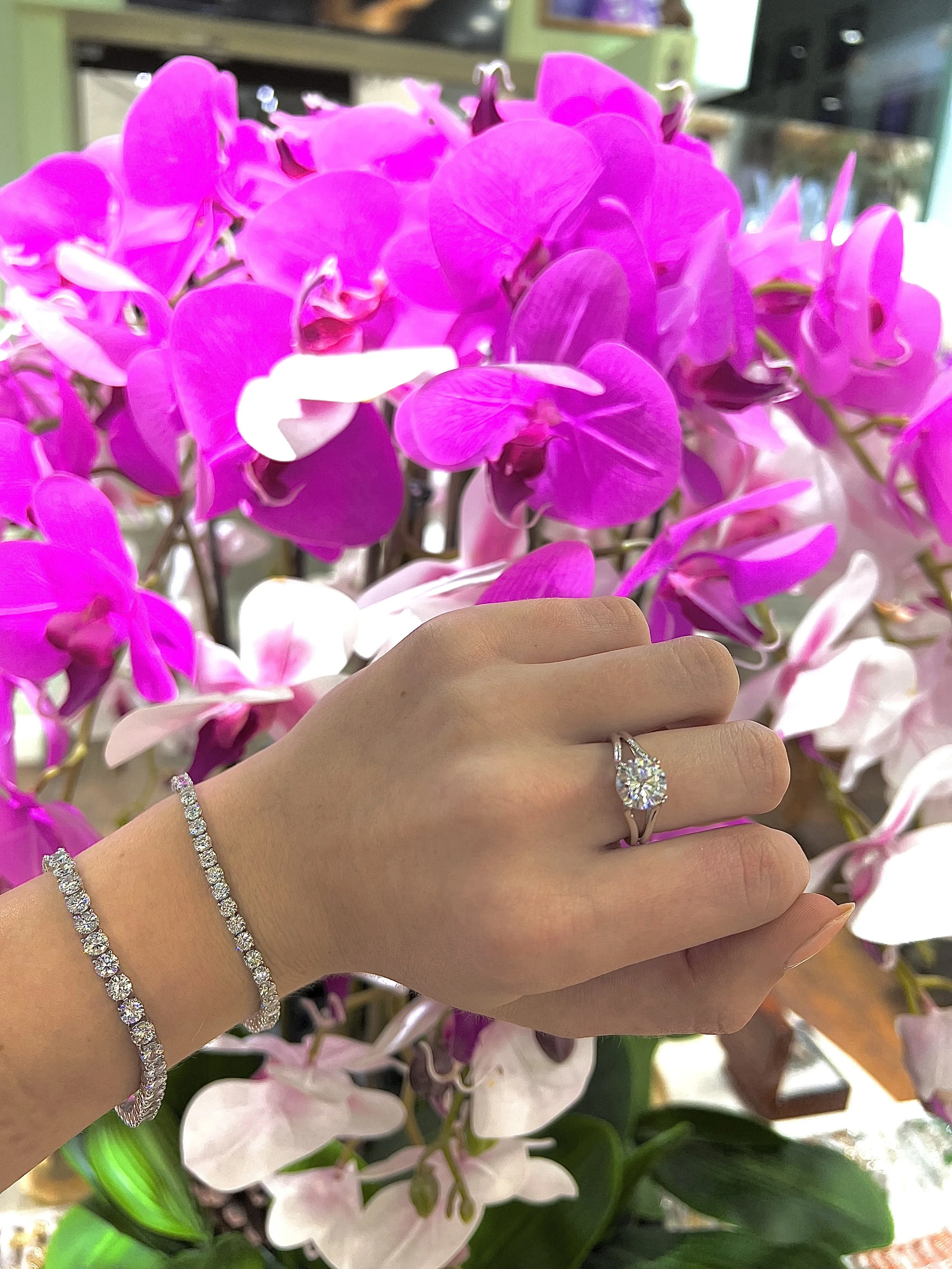 Close-up of a hand wearing a diamond engagement ring and diamond bracelets, positioned in front of a cluster of pink and white orchids.
