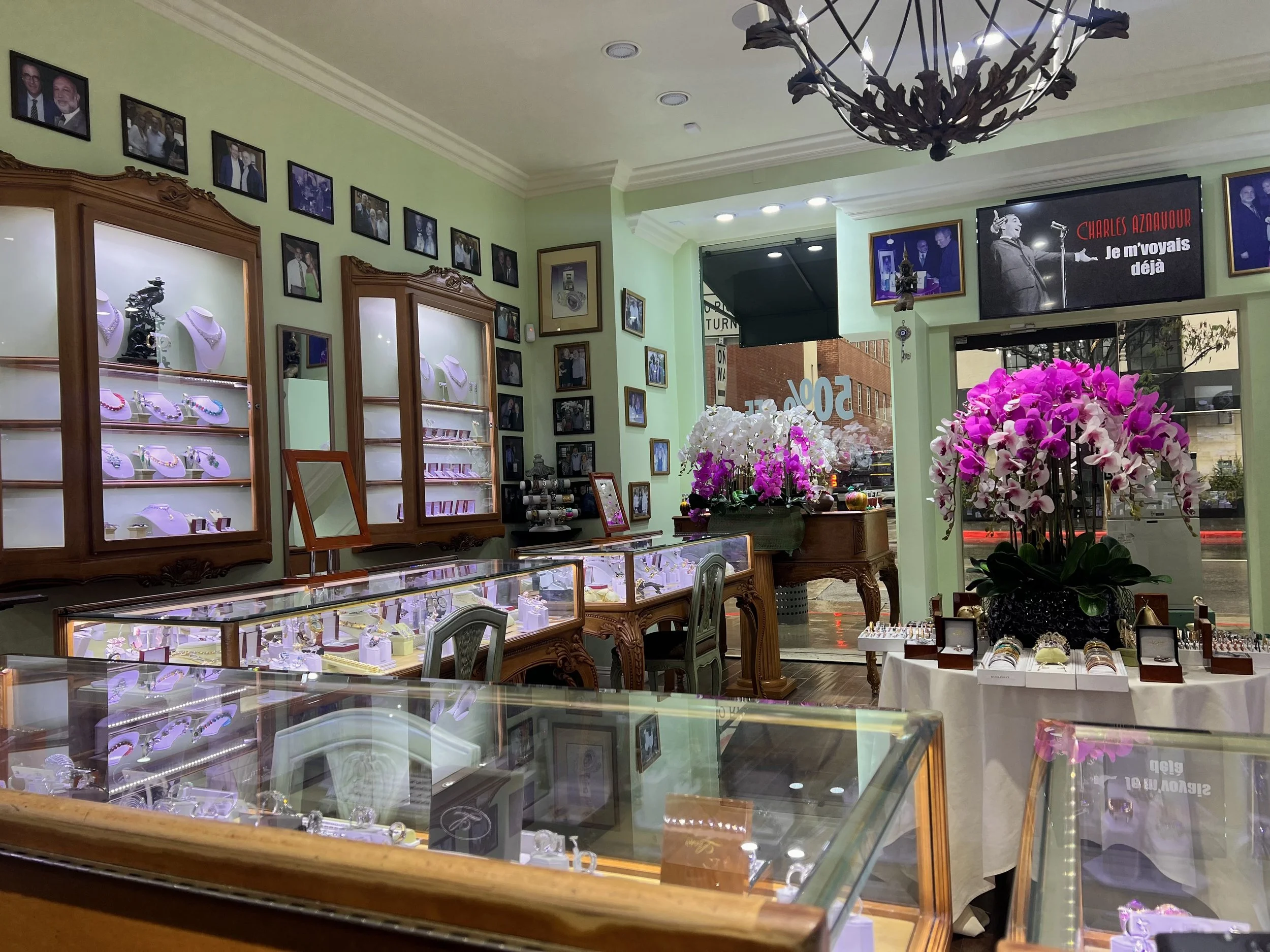 Inside a jewelry store with display cases filled with jewelry, framed photographs on green walls, and large orchid flowers on a table near the window.