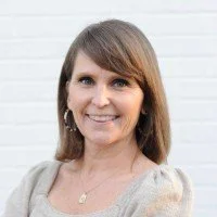 Smiling woman with brown hair, wearing earrings and a light-colored top, standing in front of a white brick wall.