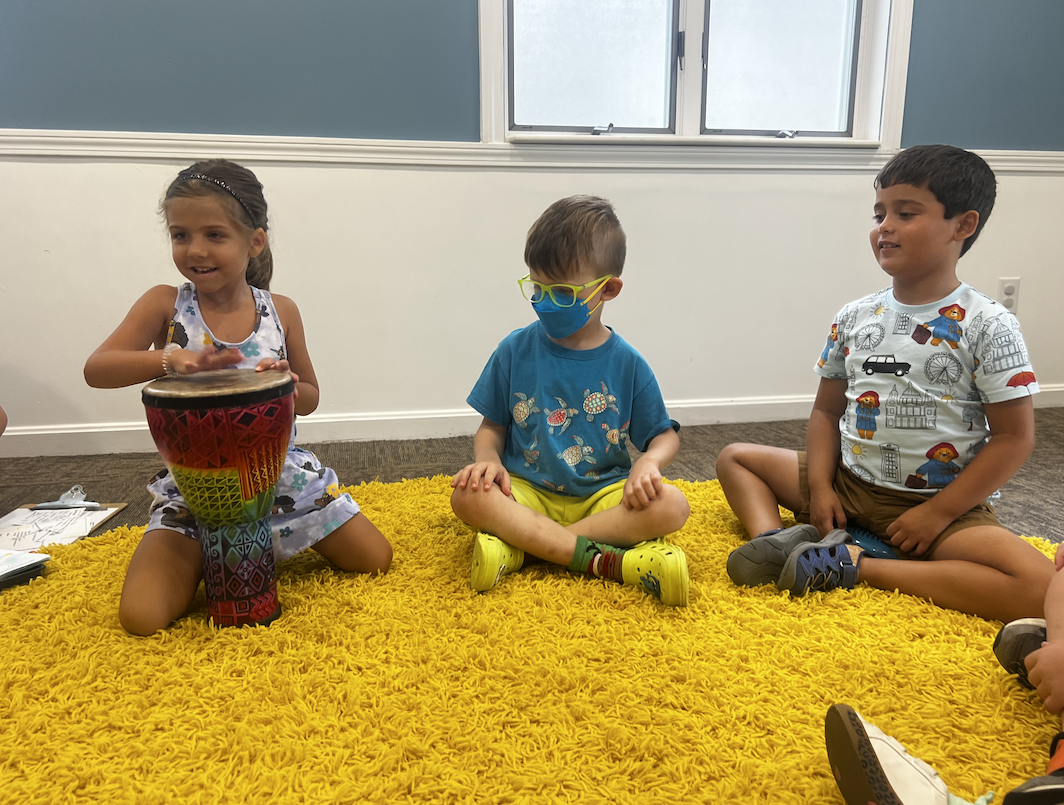 Children sitting on the yellow rug in a music therapy session at The Sonatina Center, one child playing a djembe drum