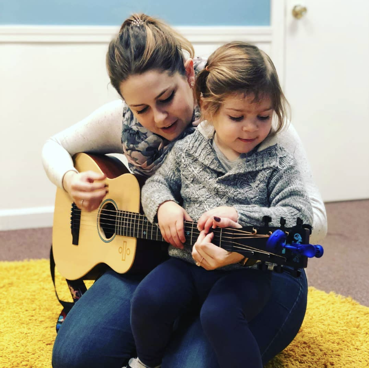Music therapist helping a young child explore the guitar during a music therapy session on the yellow rug at The Sonatina Center