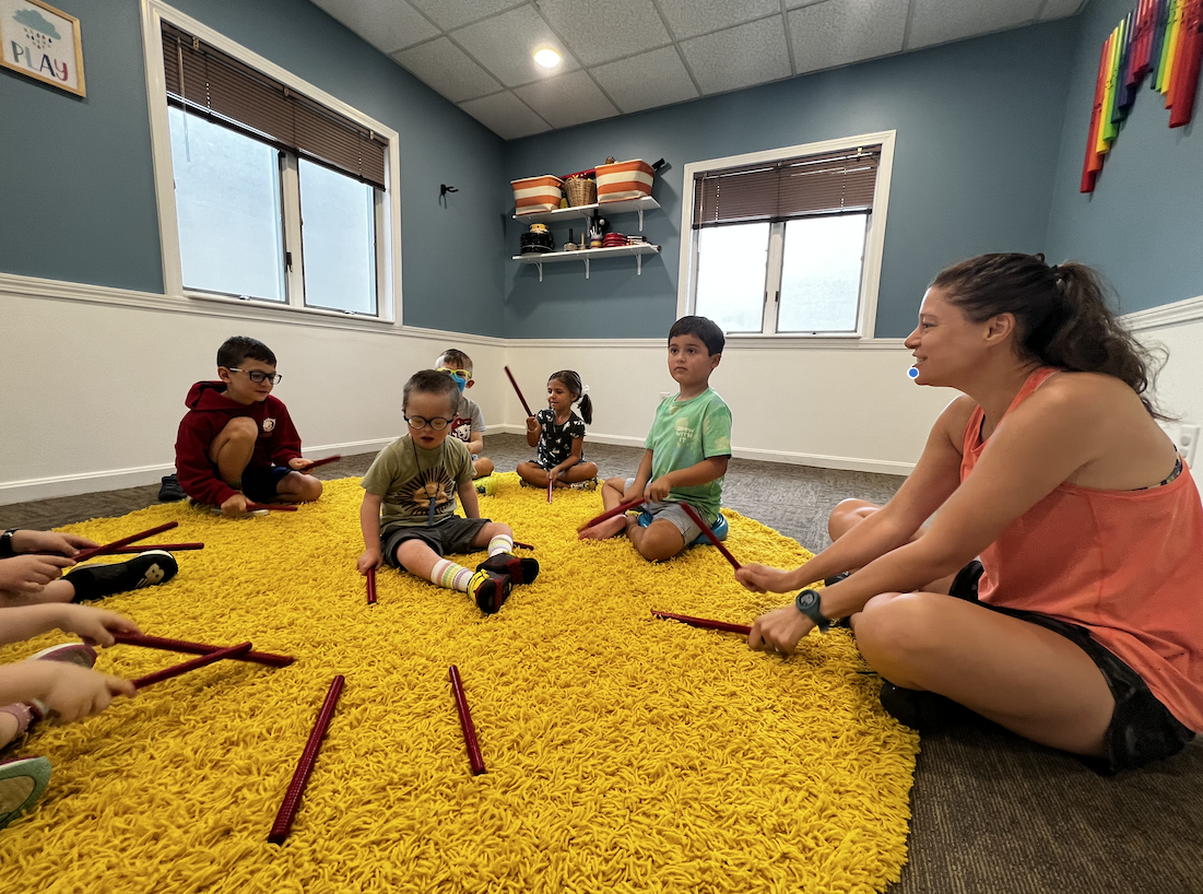 Children and music therapist sitting in a circle with rhythm sticks during a music therapy group session at The Sonatina Center