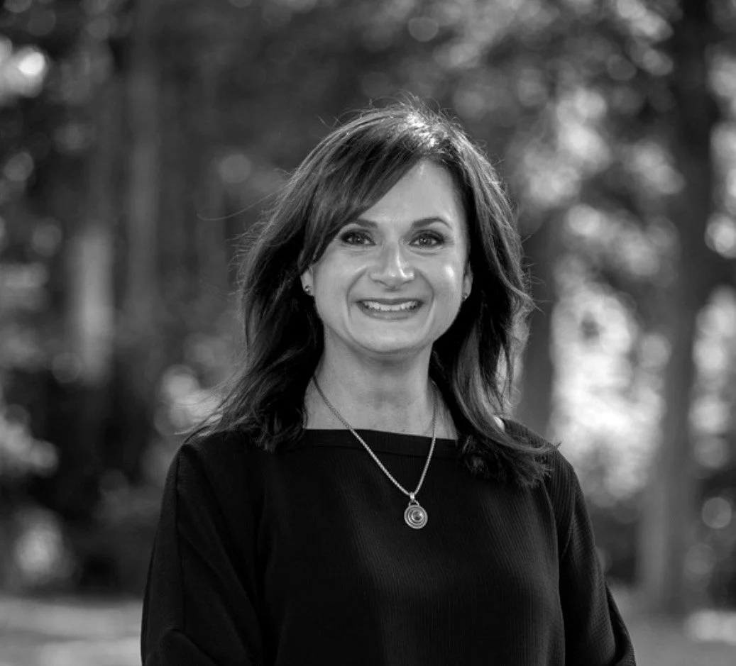 Black-and-white portrait of a smiling woman with shoulder-length dark hair, wearing a dark top and a necklace, outdoors with blurred trees in the background.