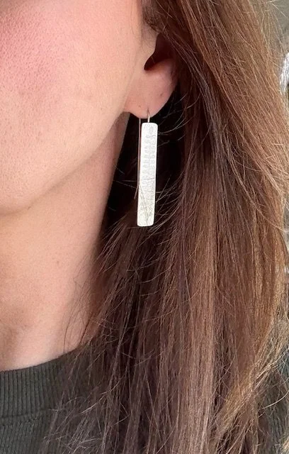 Woman wearing long rectangular silver earrings with etched botanical patterns, styled with loose hair