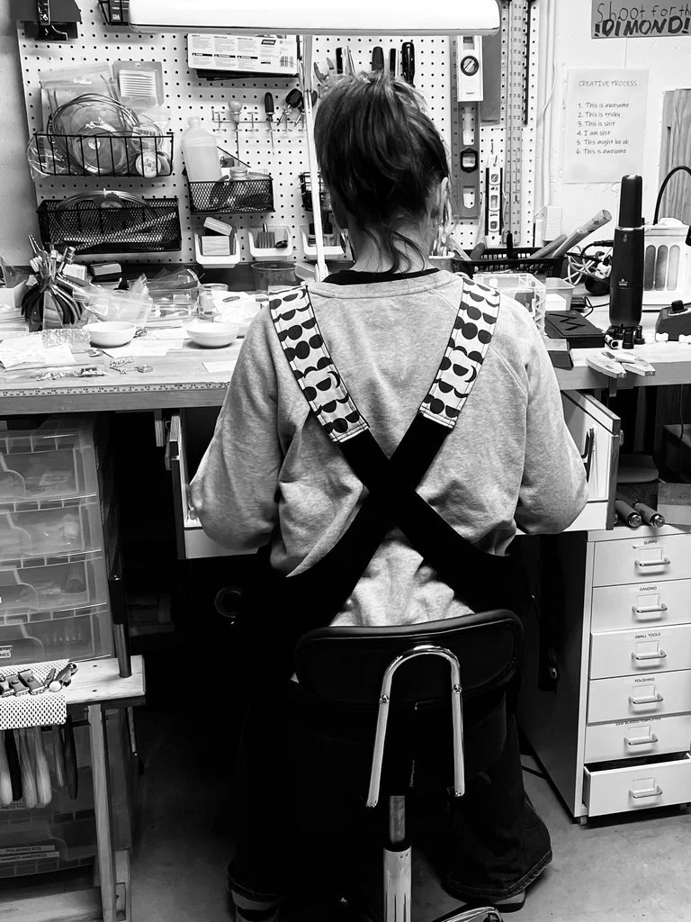 Kelly of KFinn designs at her workbench in a workshop, making handcrafted jewelry surrounded by tools and supplies, photographed from behind.
