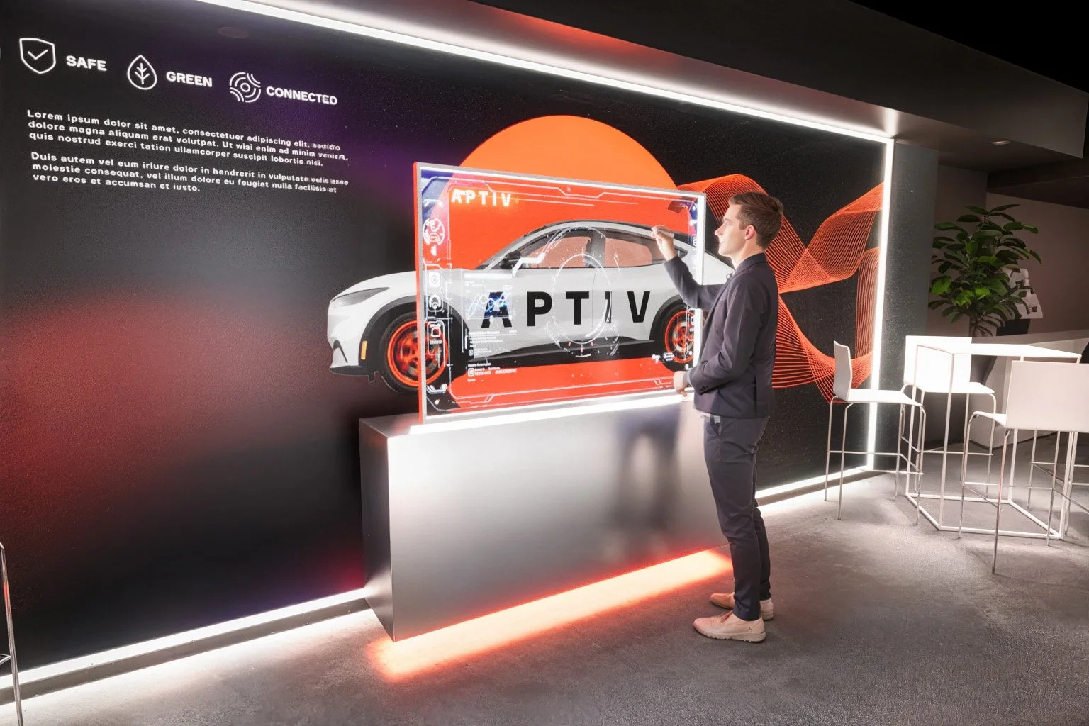 Man in dark suit interacts with a transparent digital display featuring a white sports car with red accents, labeled 'APTIV', mounted on a stand in a modern office or showroom with chairs, a table, and a large digital screen with placeholder text in the background.