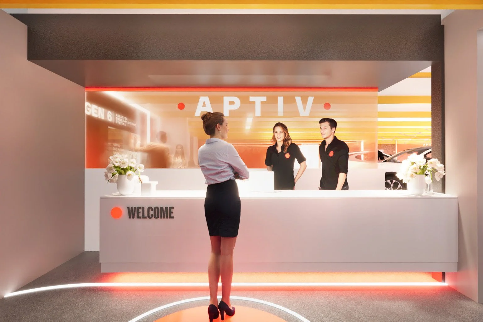 Reception area with a white desk labeled 'WELCOME' and a woman standing in front of it, talking to two staff members behind the counter. The background features a sign with 'APTIV' and modern decor with flowers and warm lighting.