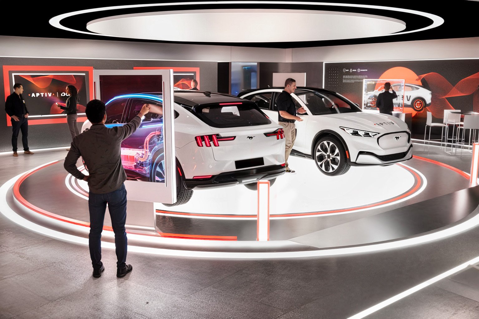 An auto show display with two white Ford vehicles on a circular platform, surrounded by a few visitors and staff. The background includes a large digital screen and promotional material, with a modern, well-lit environment.
