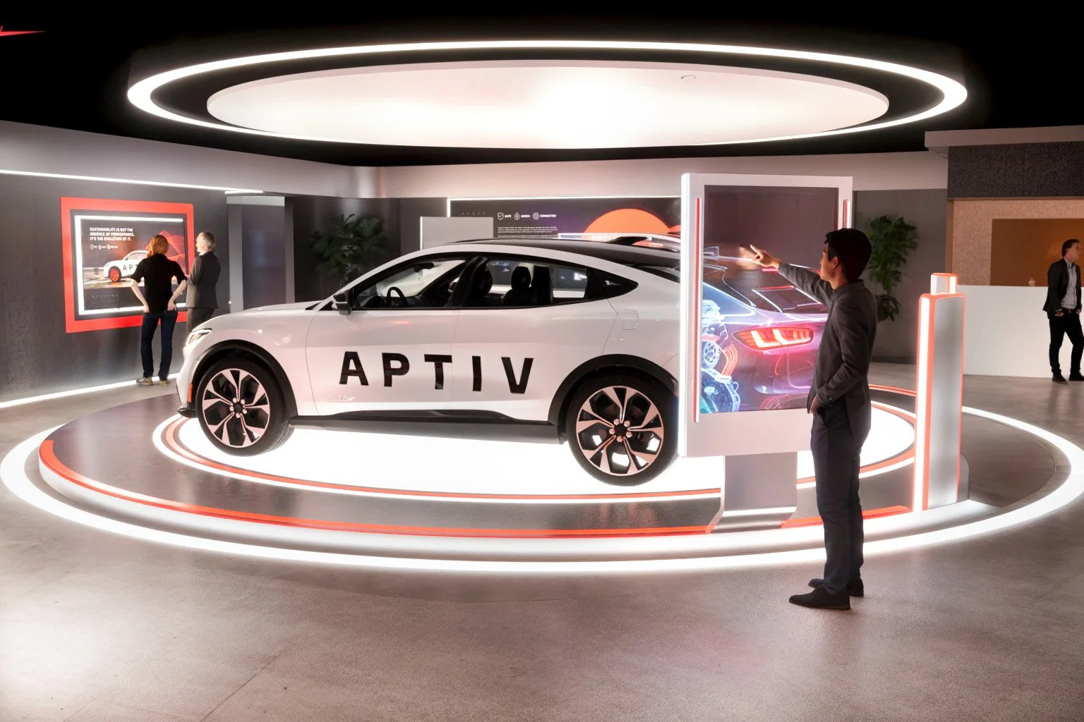 Showroom with a white car labeled 'APTIV' on a circular platform, surrounded by digital displays and people observing and interacting with the exhibits.