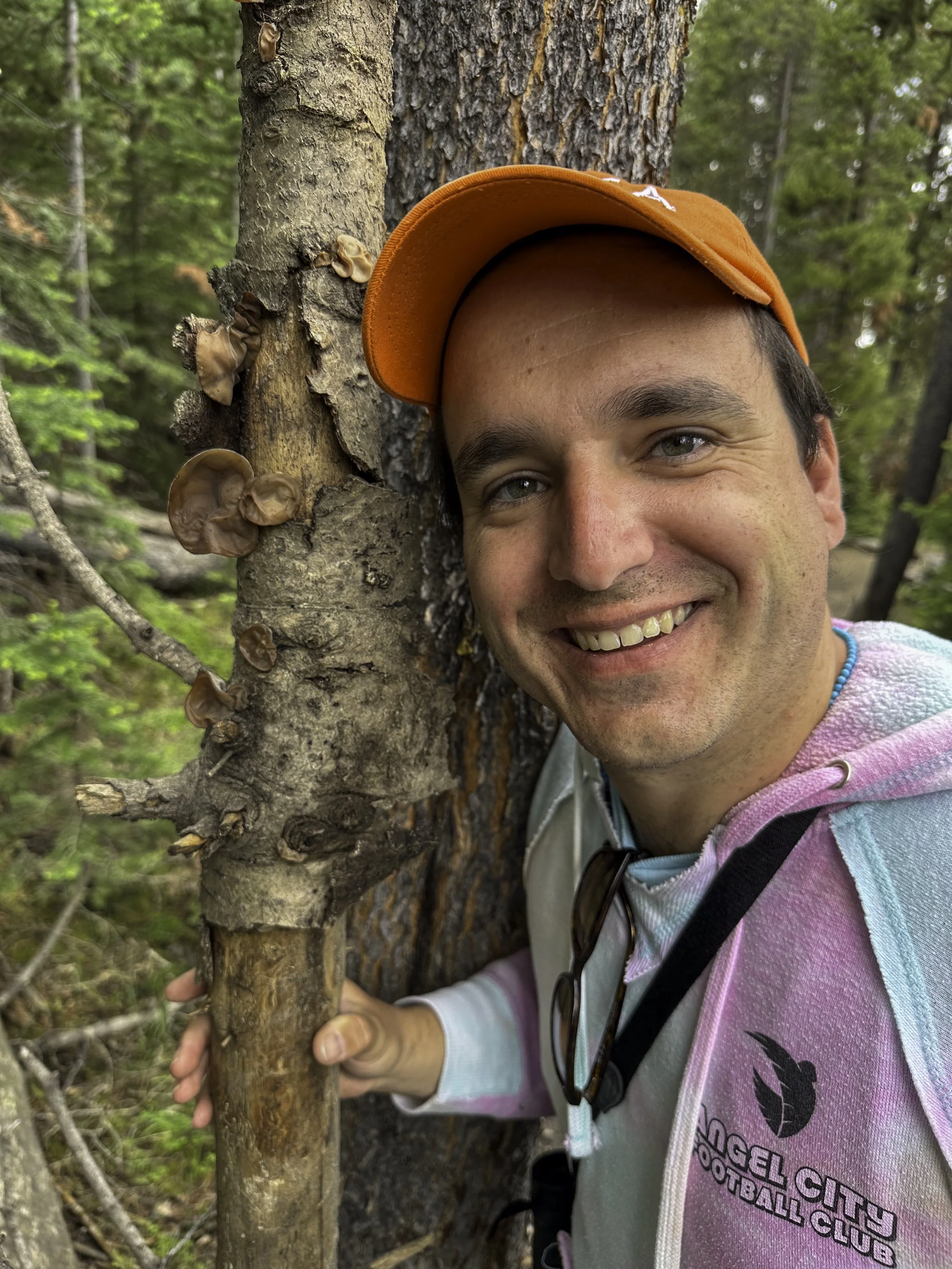 A person smiling and hugging a tree in a forest, wearing an orange cap and a hoodie with the logo of the Engelwood City Football Club.