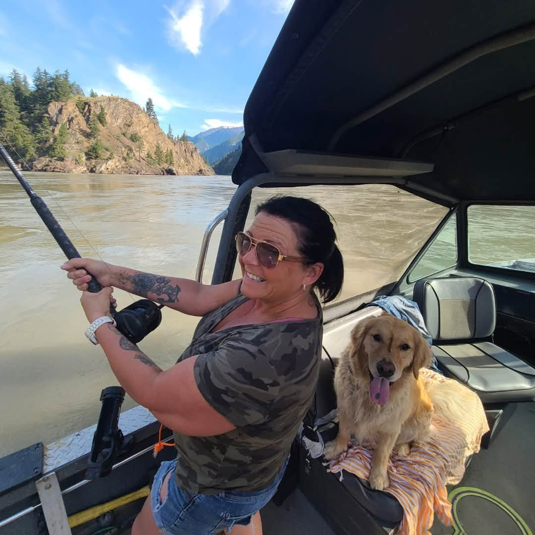 Woman fishing from a boat on a river with a dog sitting beside her, mountain landscape in the background, clear sky, woman wearing sunglasses, camouflage t-shirt, and denim shorts.