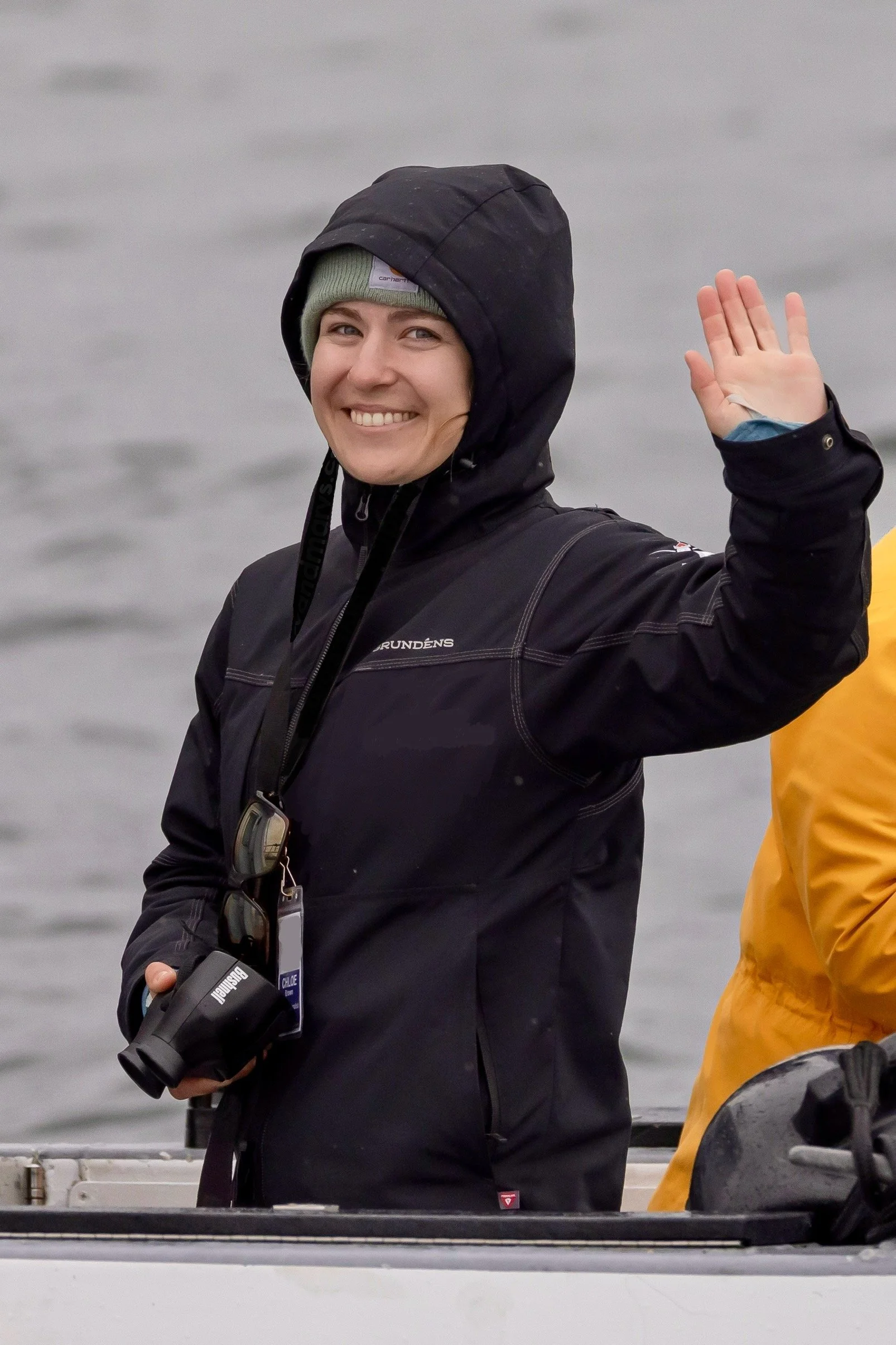 A woman in a black waterproof jacket waving and smiling, standing on a boat with a body of water in the background.