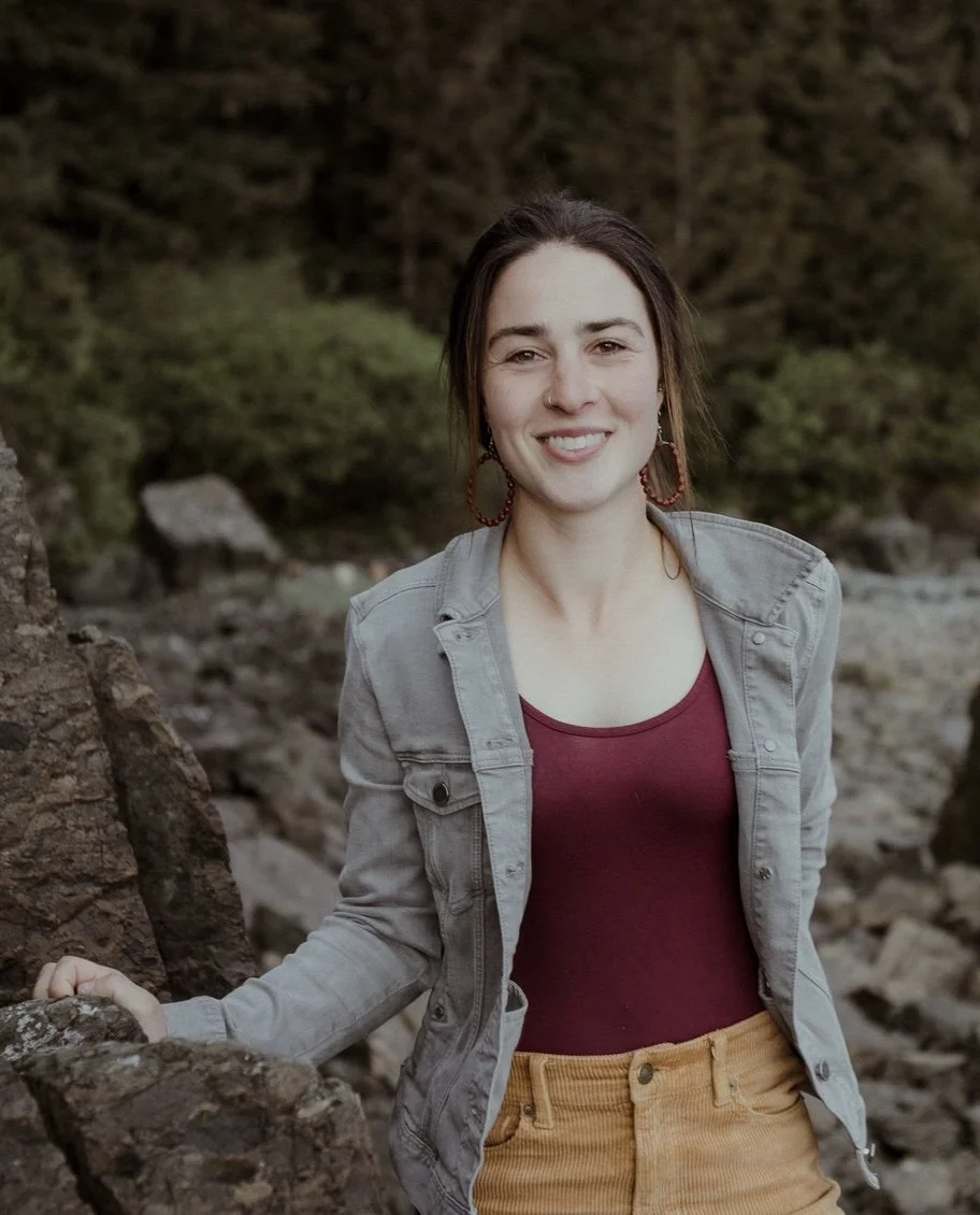 A young woman outdoors standing among rocks with a forest background, smiling, wearing a gray jacket, maroon top, and mustard yellow pants.