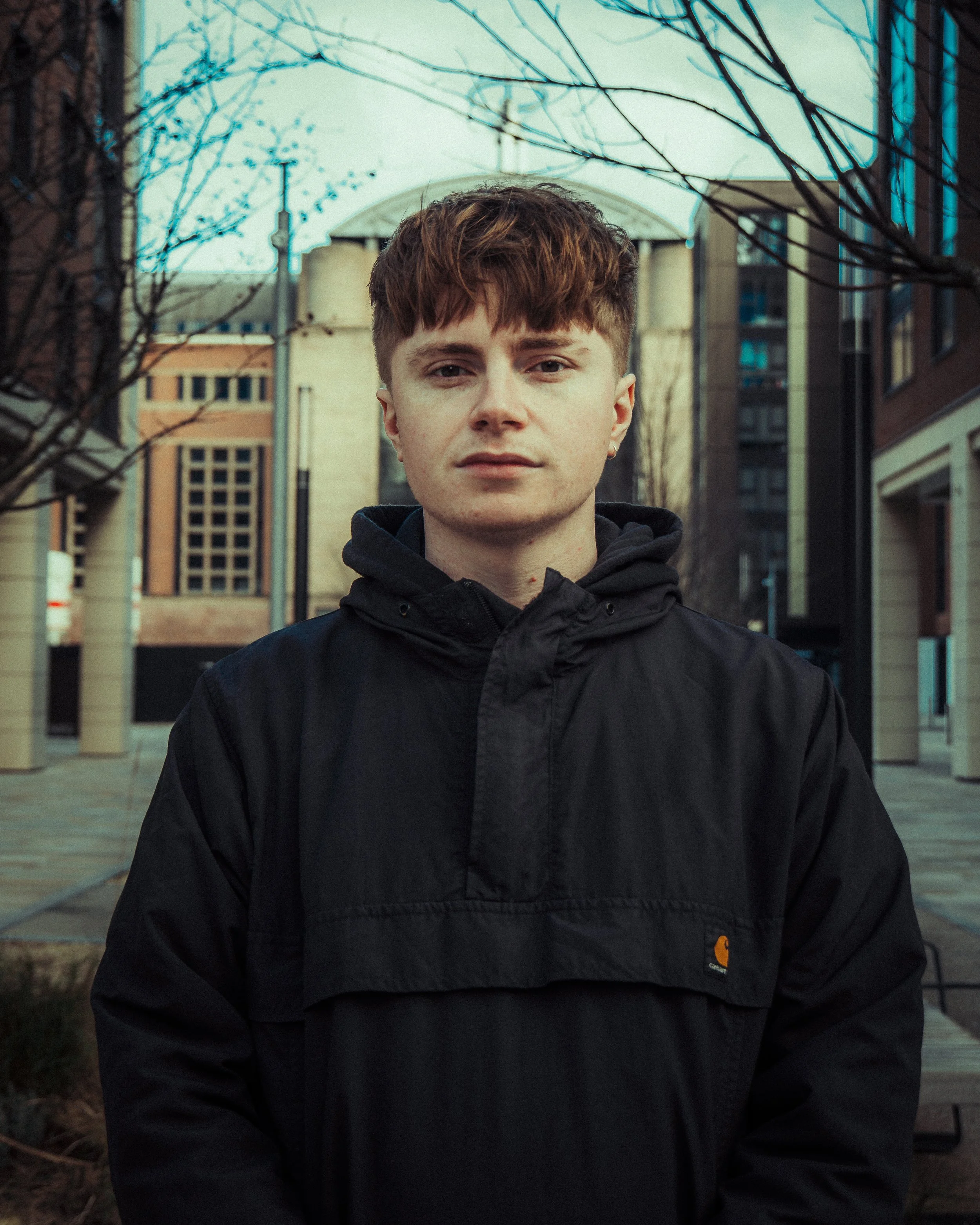Young man with brown hair wearing a black Carhartt jacket standing outdoors in an urban setting with buildings, trees, and an overcast sky behind him.
