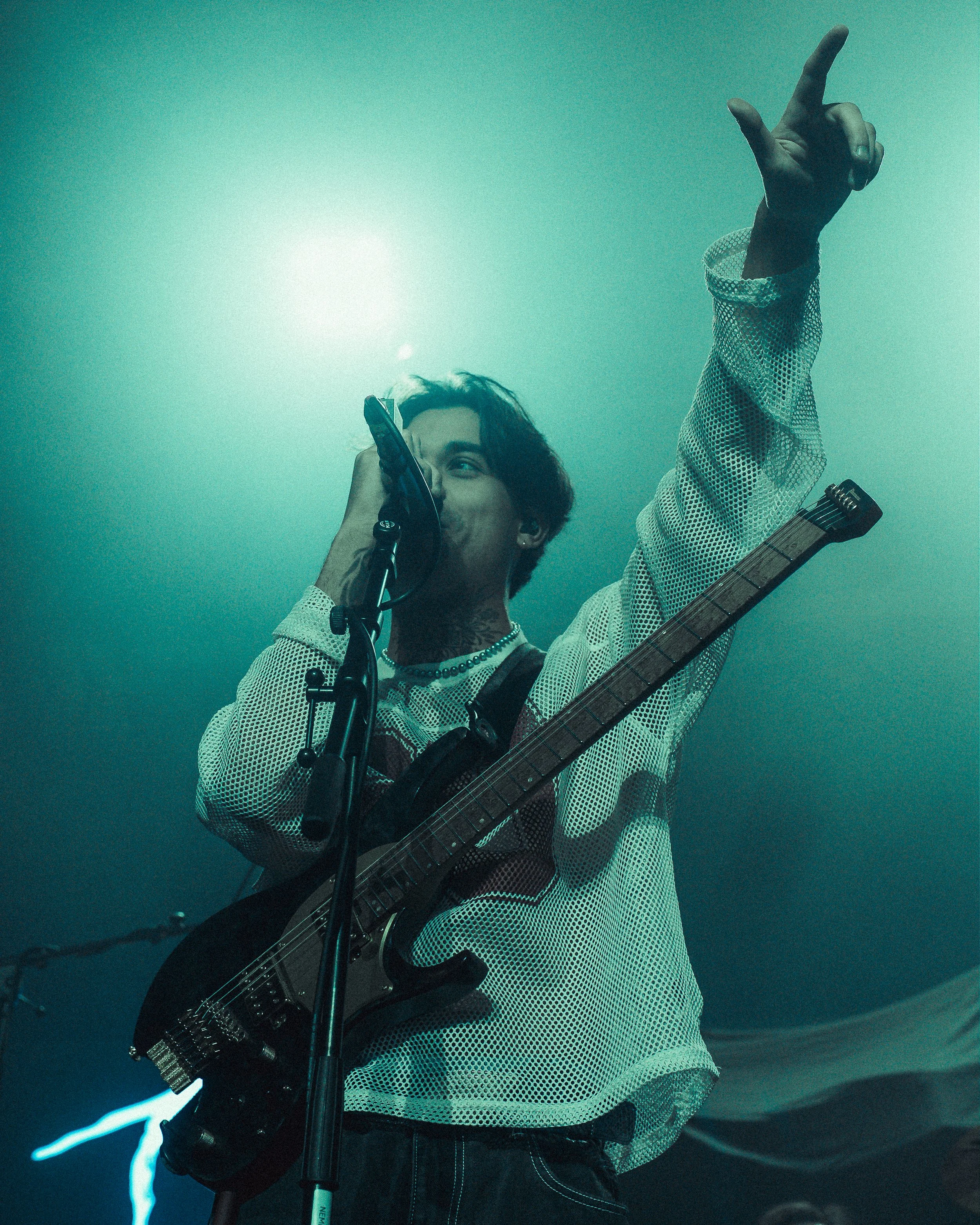 A musician with dark hair performing on stage, singing into a microphone while holding a guitar, with his right hand raised and pointing upwards.