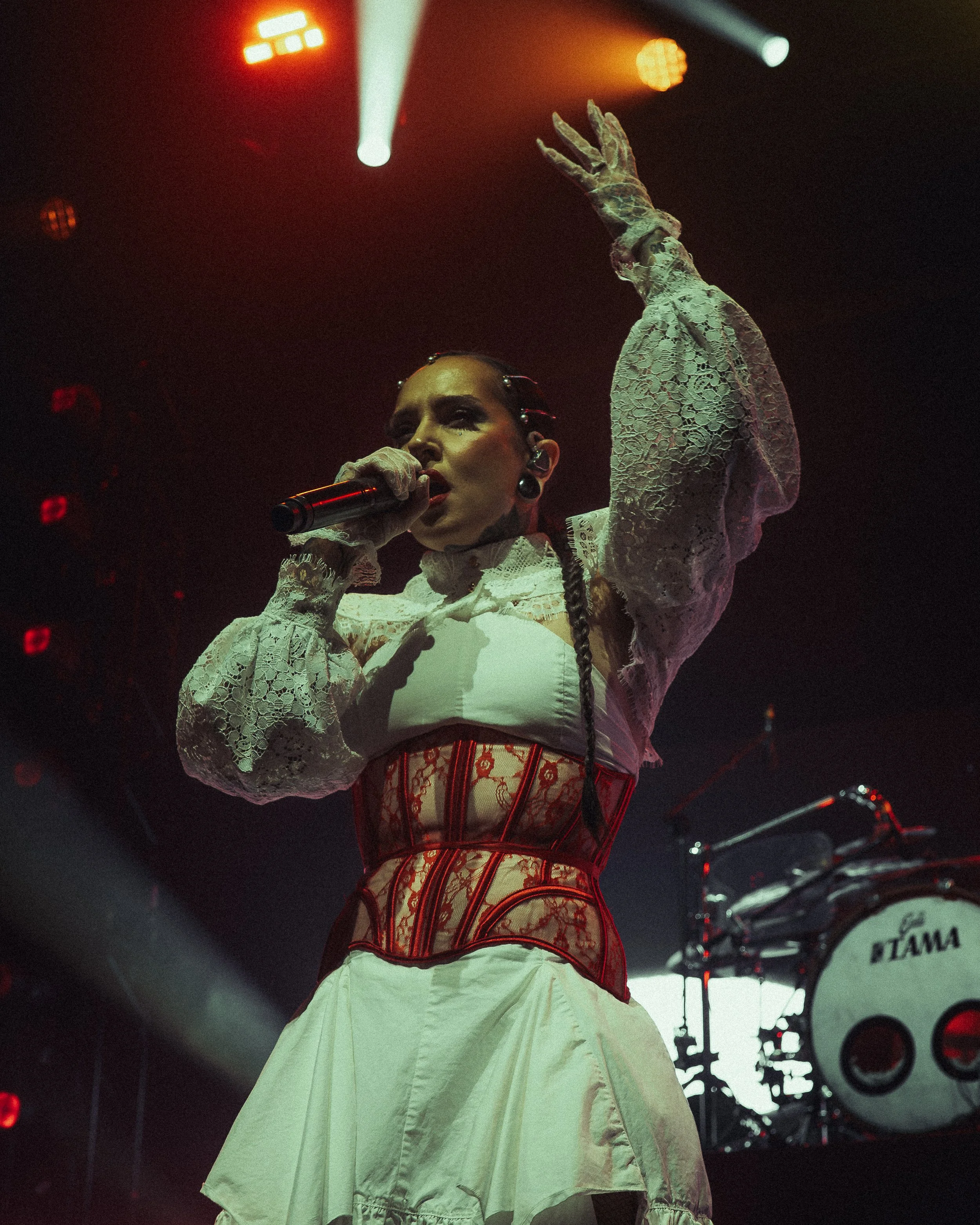 A female singer performing on stage under colorful lights, wearing a lace blouse with puffy sleeves, a red corset, and a cream-colored skirt.