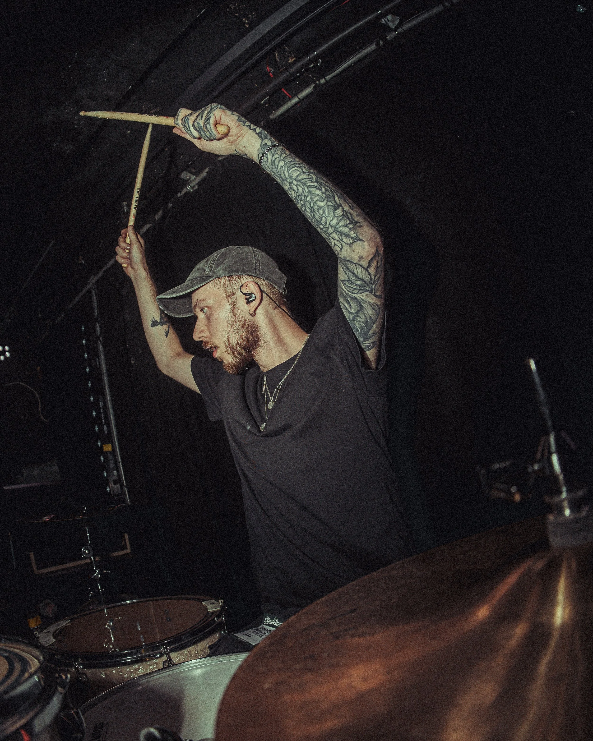 A man wearing a cap and black t-shirt, with tattoos on his arms, is playing drums at a dark venue. He is holding drumsticks above his head.