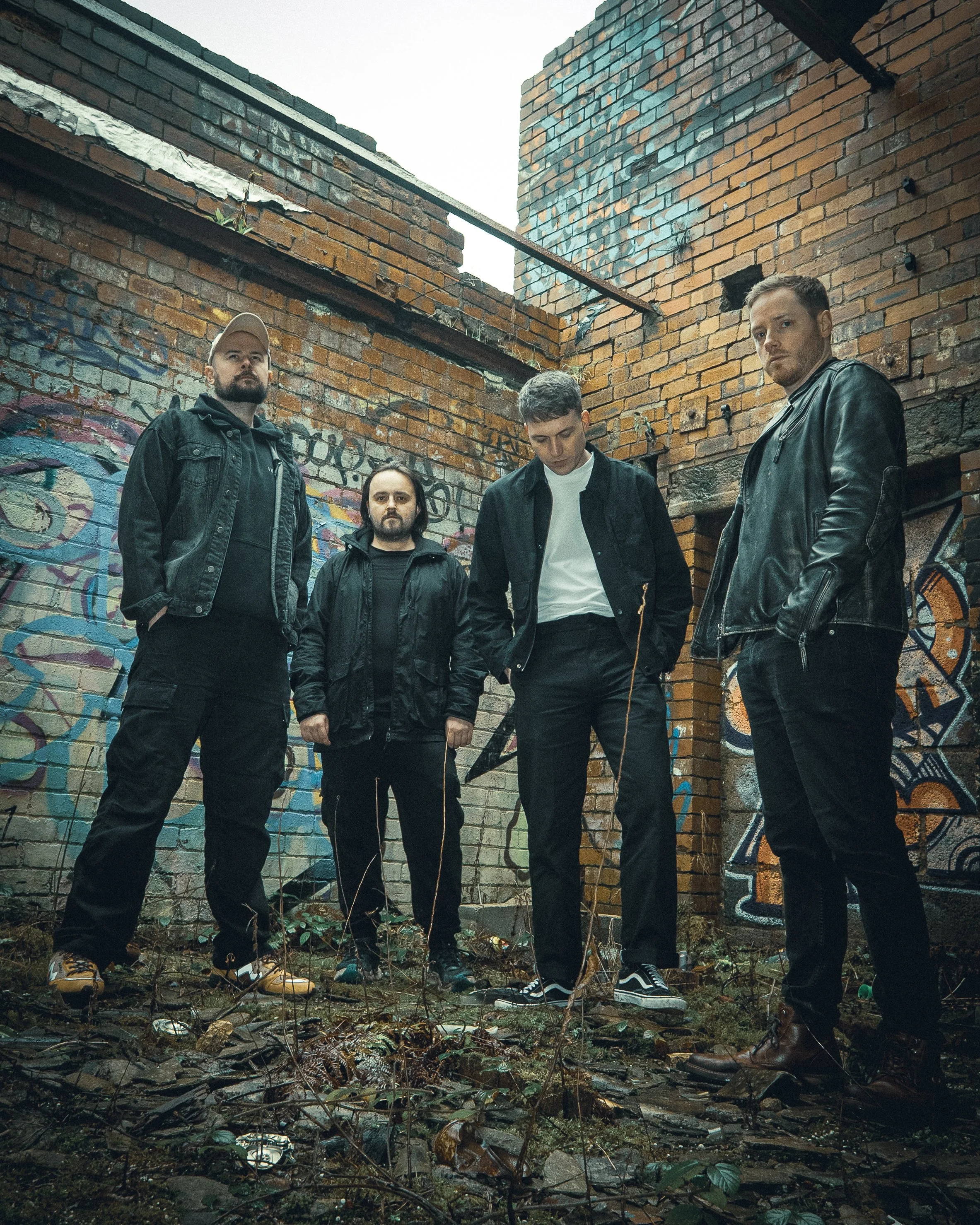 A group of five young men standing in an alleyway with graffiti-covered brick walls. They are dressed in dark clothing and have serious expressions.