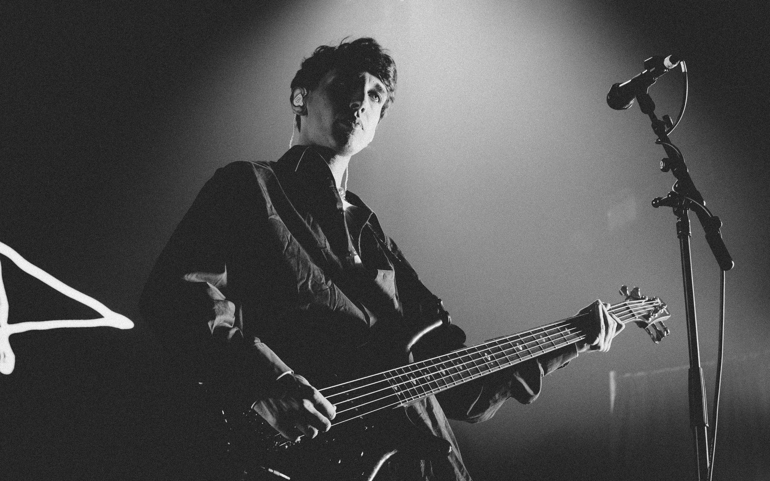 A man holding a bass guitar and standing near a microphone on stage, illuminated by stage lights in a concert setting.