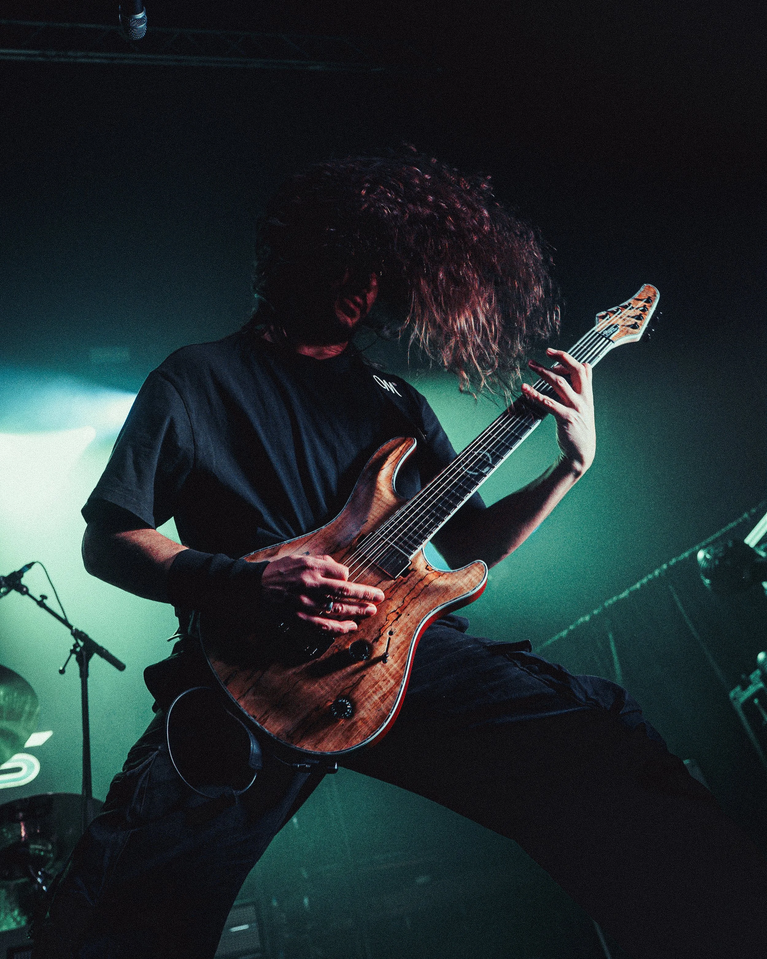 A musician with long, curly hair playing an electric guitar on stage under green and blue stage lighting.