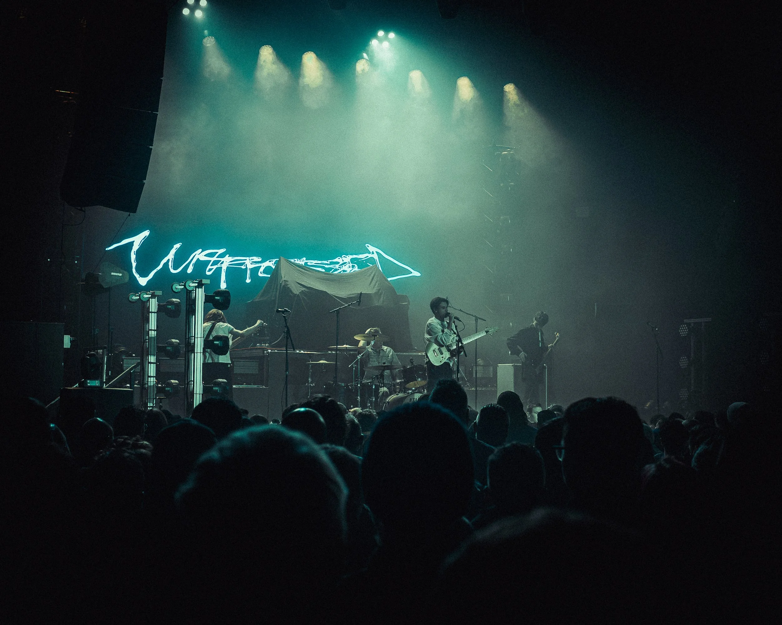 A band performs on stage with a large crowd watching. Stage lights are shining down on the musicians, and a neon sign or projection appears in the background.