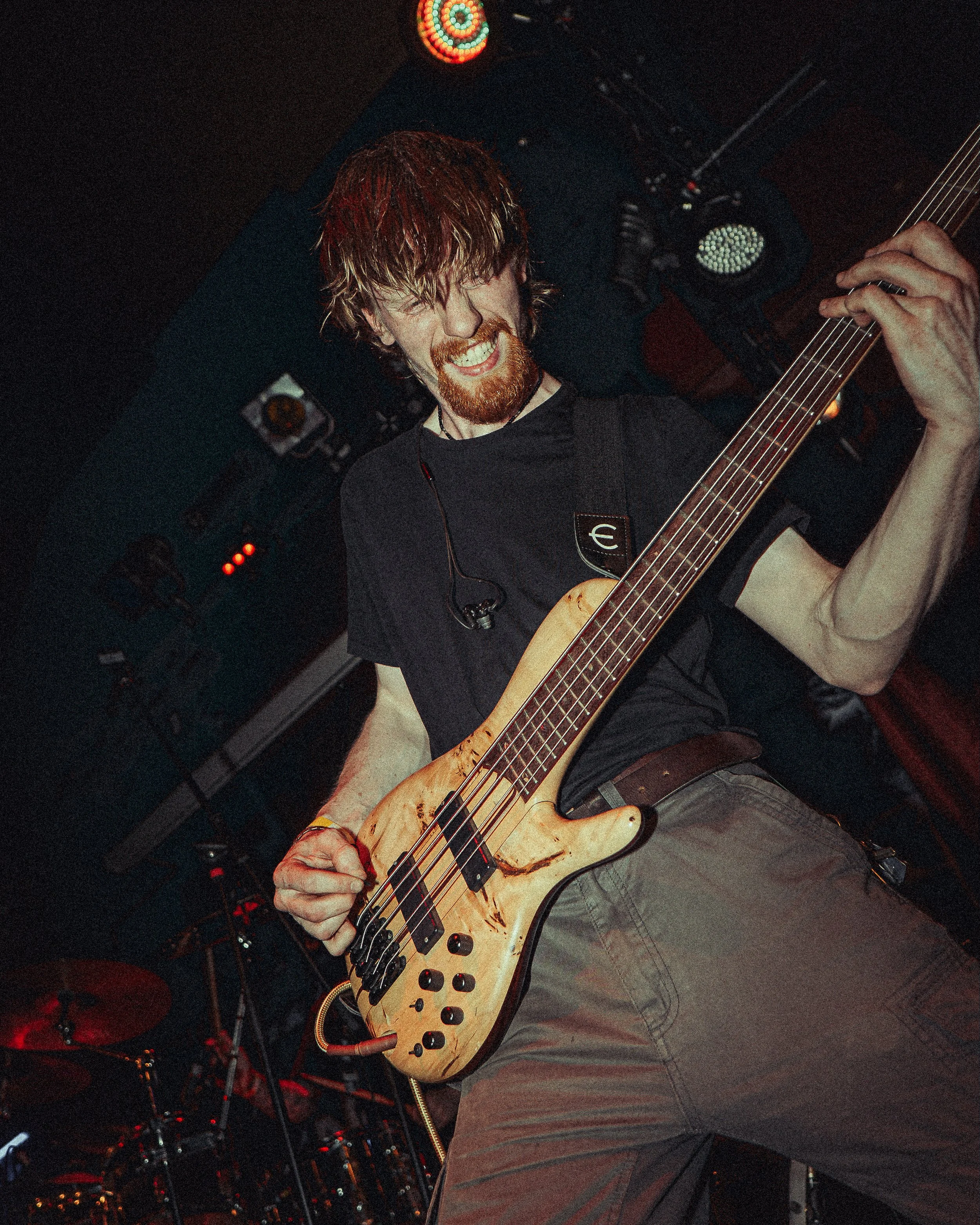 A musician playing an electric bass guitar during a live performance, smiling and wearing a black t-shirt. The background includes stage lights and drum set.