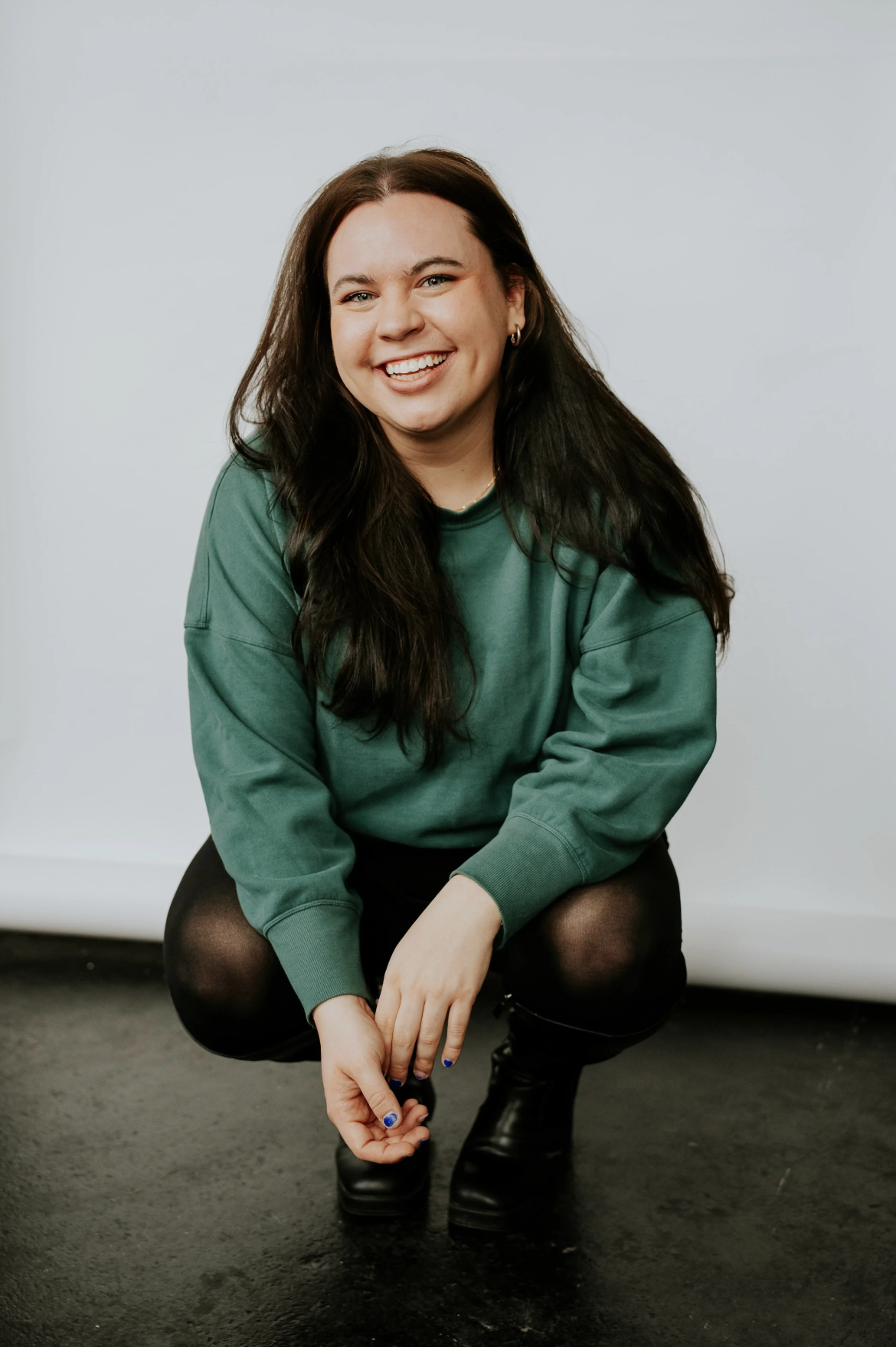 Young woman with long dark hair, wearing a green sweatshirt, black pants, and black boots, sitting on the floor, smiling at the camera against a plain background.