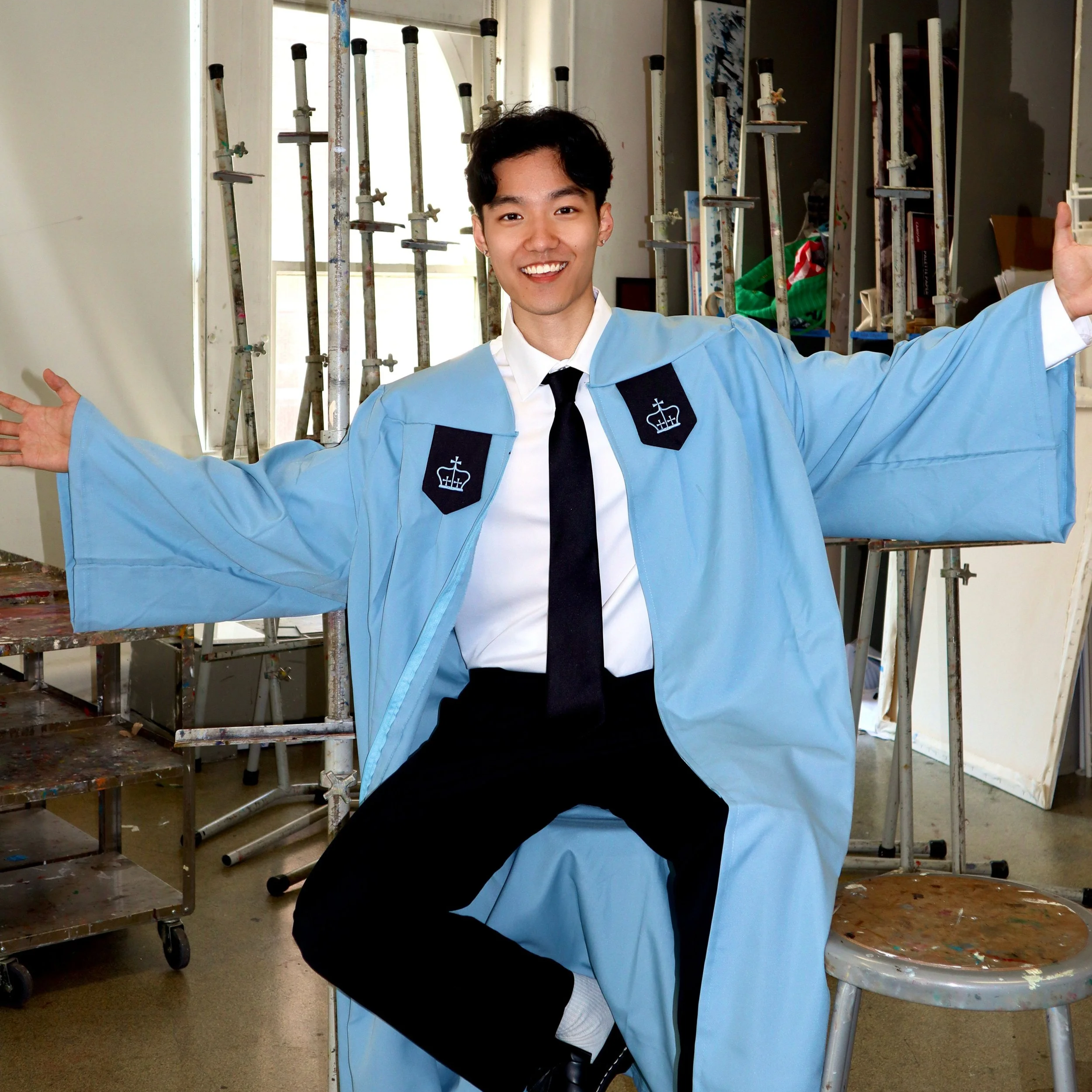 A young man in a blue graduation gown and black tie smiling with arms outstretched in a room filled with painting supplies and scaffolding.