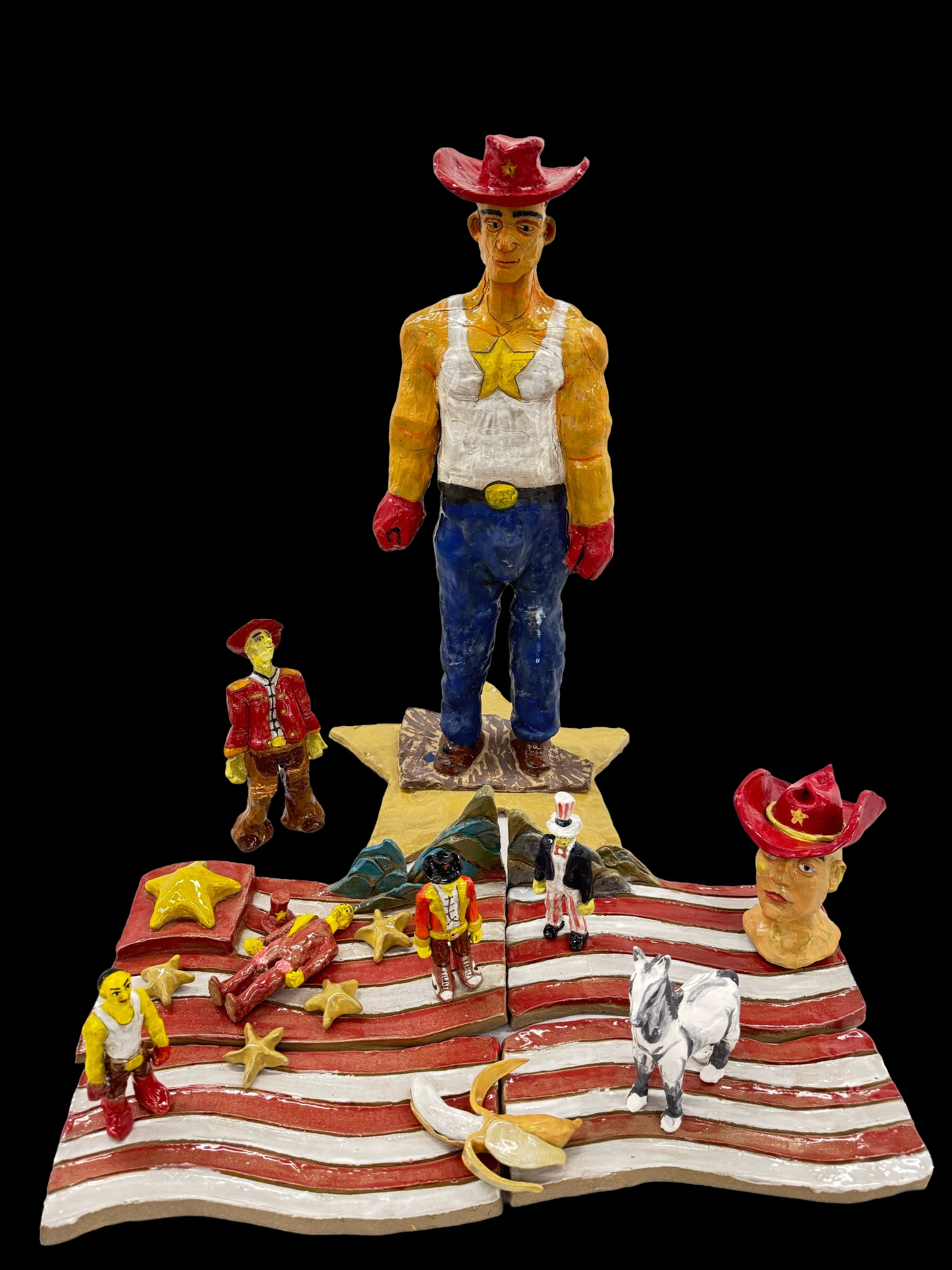 Colorful handcrafted diorama of a cowboy scene with a large cowboy figure, smaller figures, animals, stars, and American flag-themed elements on a textured base.