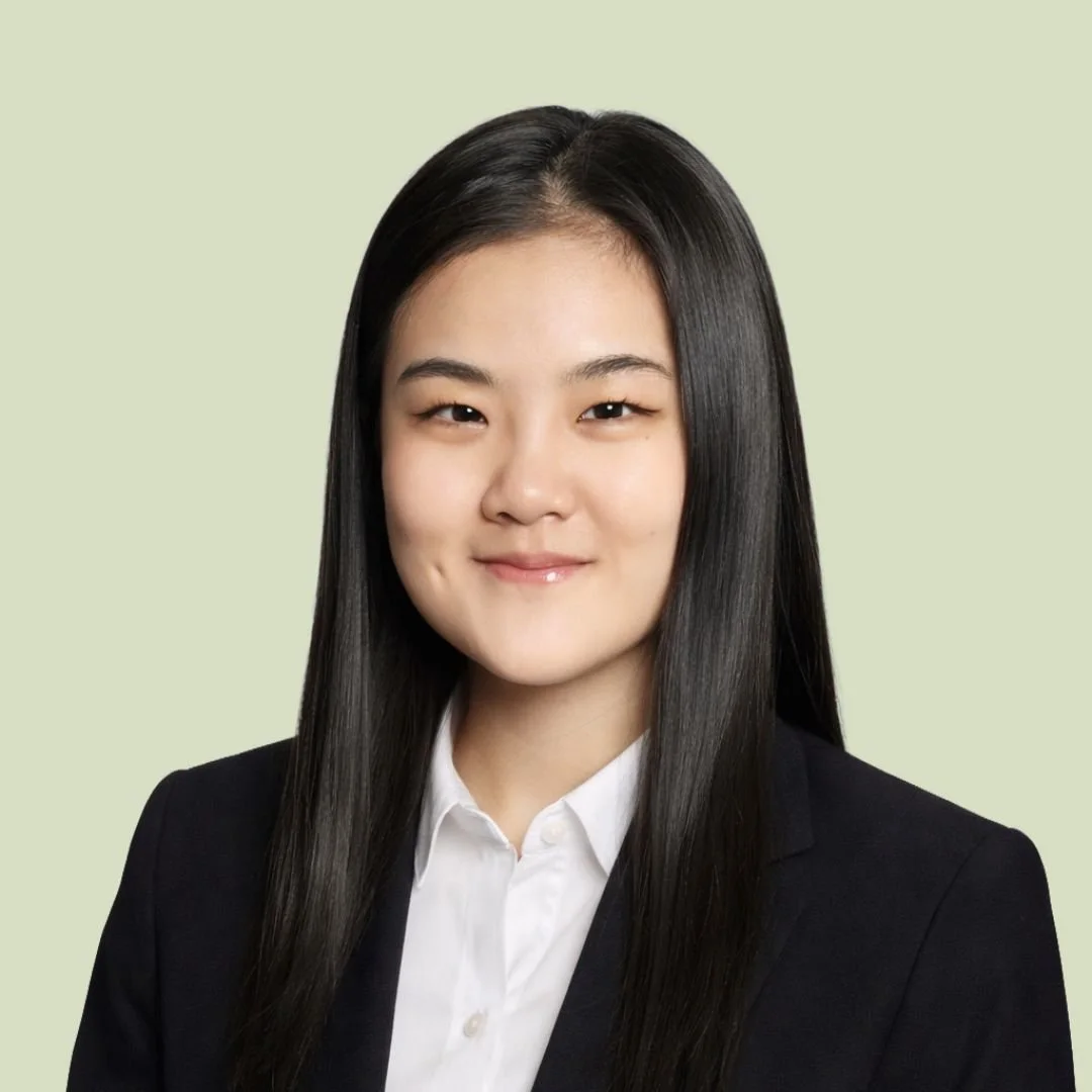 Professional headshot of a young woman with straight black hair, wearing a black blazer over a white collared shirt, smiling gently against a light green background.