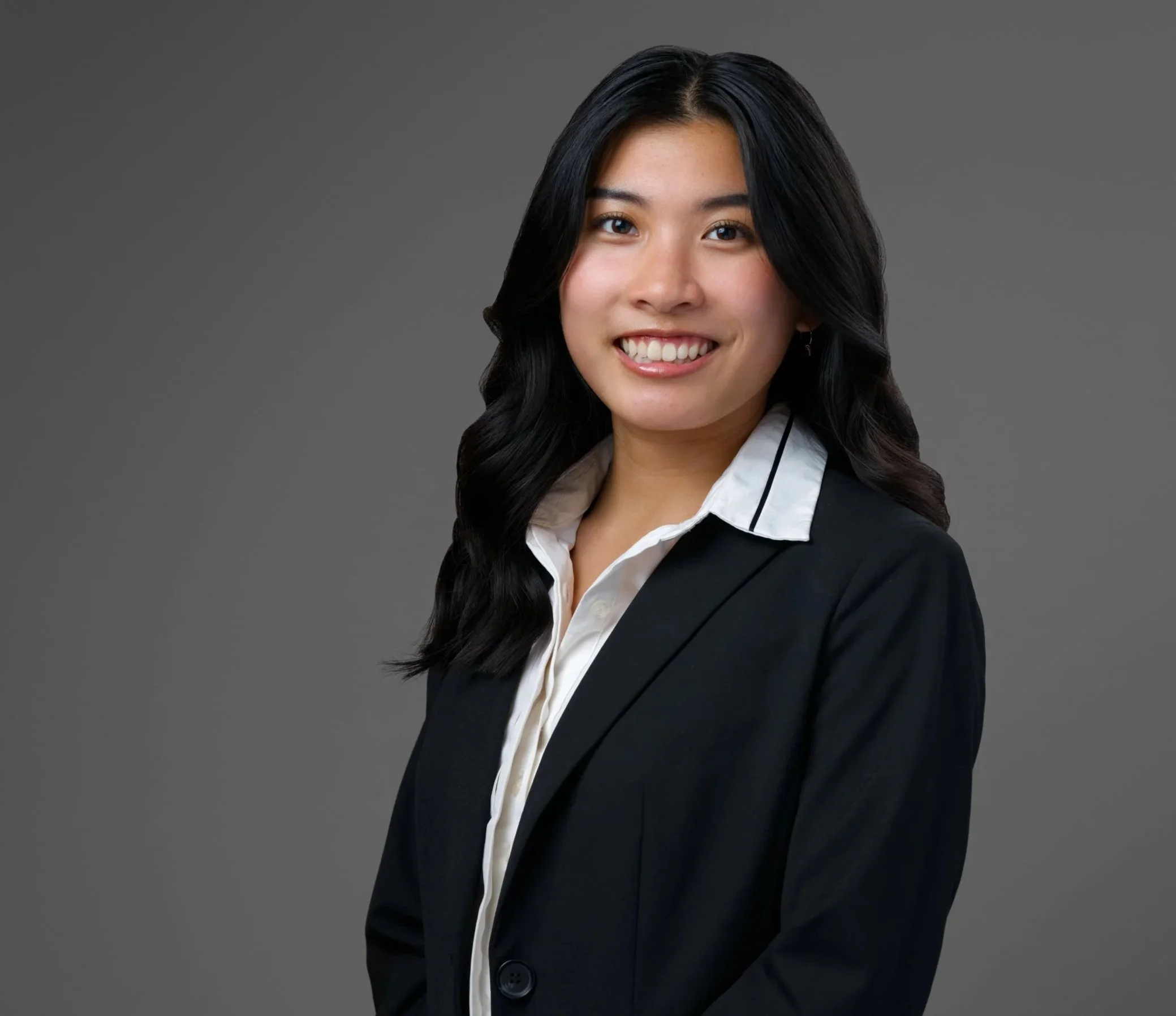 A young Asian woman smiling wearing a blazer and white collared shirt against a plain gray background.