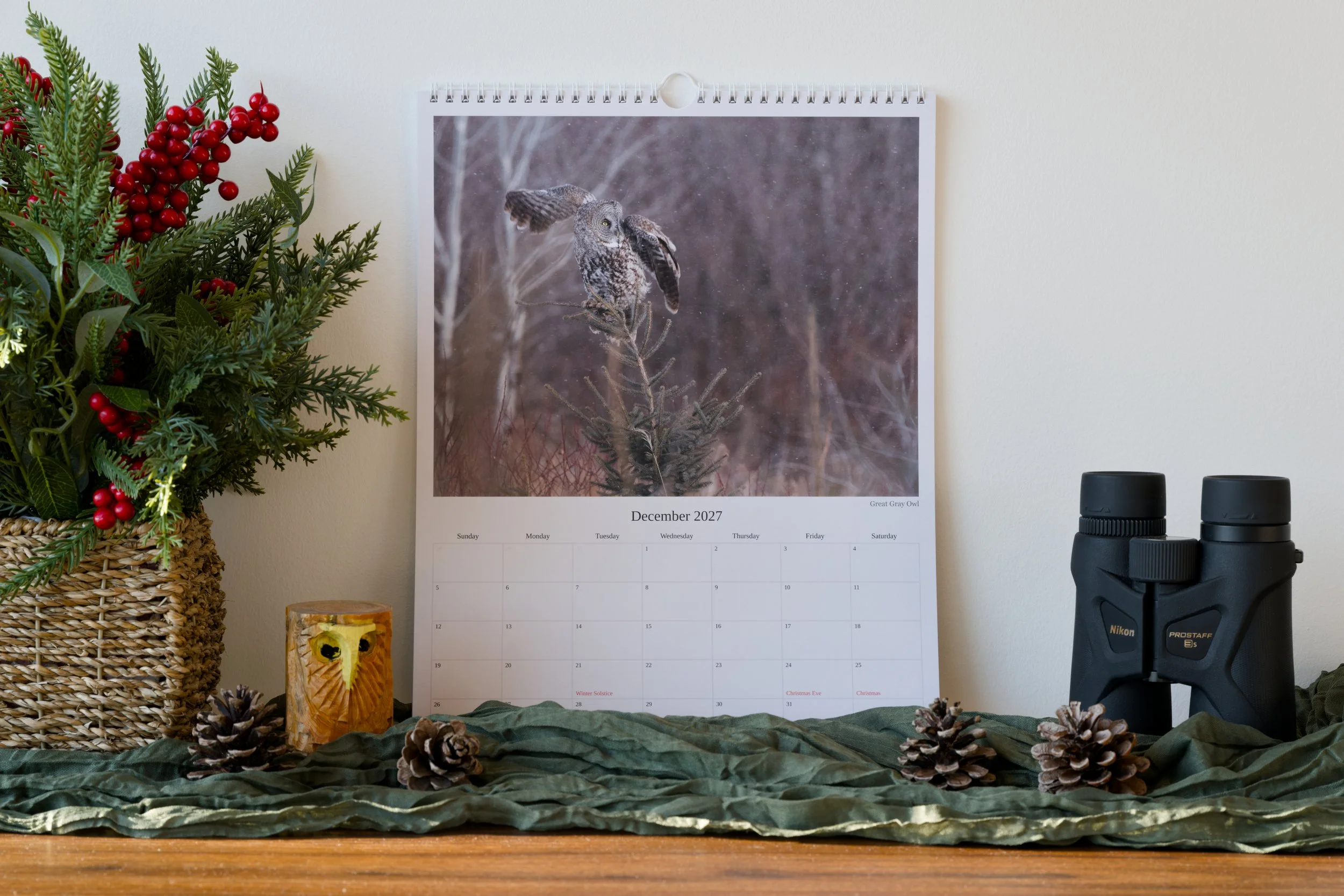 A calendar depicting a Great Gray Owl is leaning against a wall while resting on a table with winter-themed decor surrounding it and binoculars on the right.