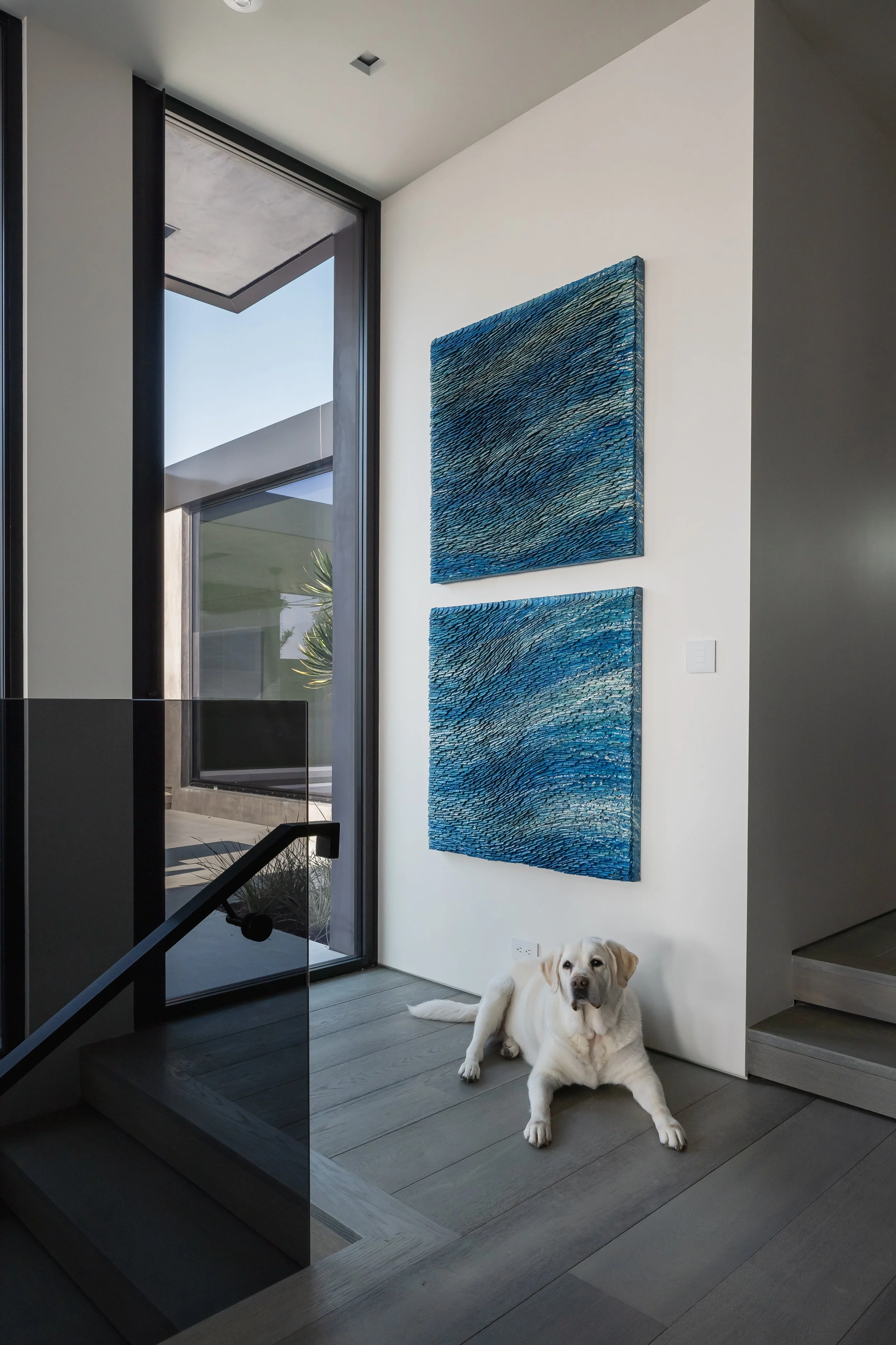 Interior view of a modern house with stairs, large window, and two blue abstract paintings on white wall; a Labrador retriever lying on the floor near staircase.