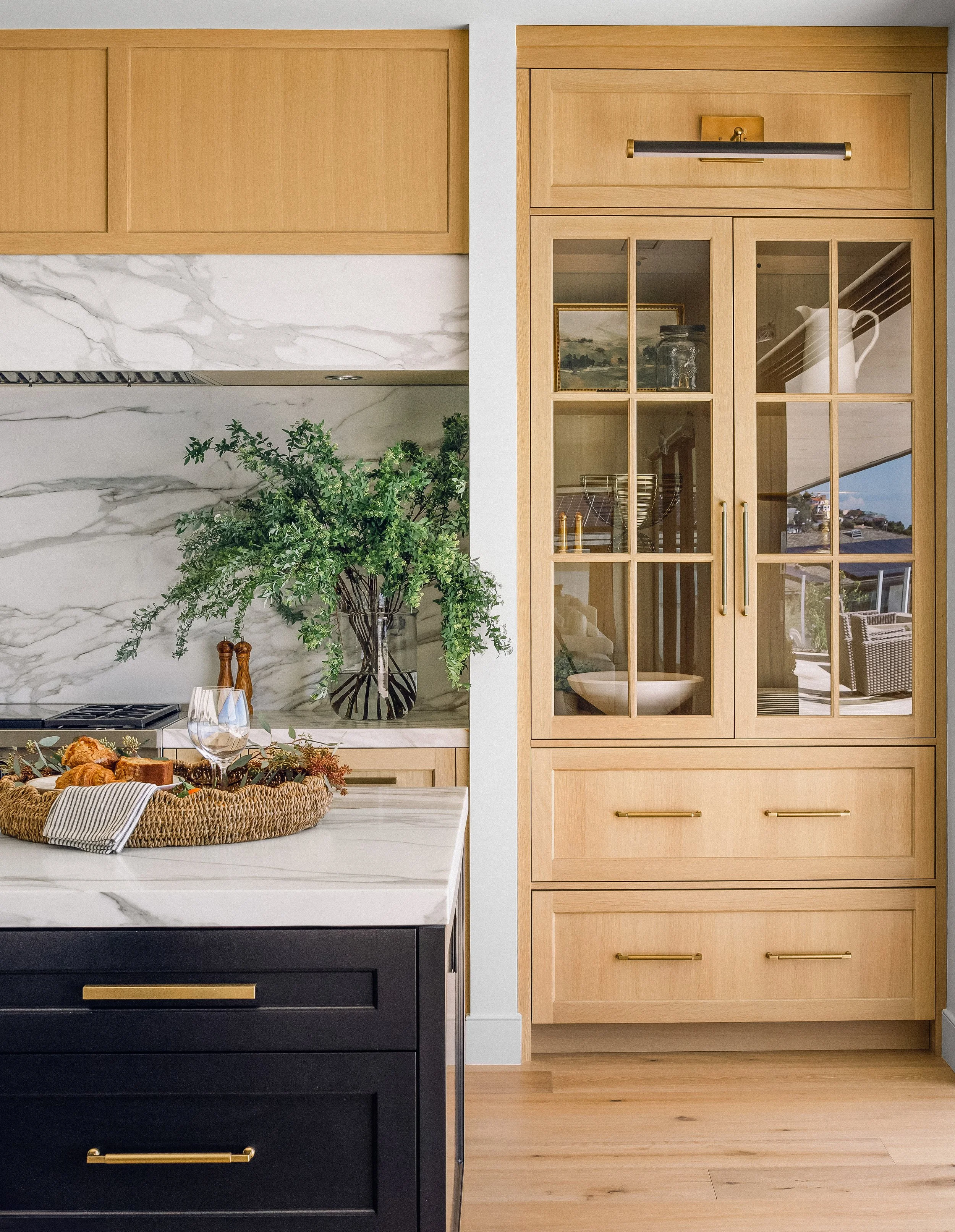 Interior view of a modern kitchen featuring a black kitchen island with a white marble countertop, a large plant in a glass vase, and a woven basket with food. To the right, there is a tall light wooden cabinet with glass-paneled doors and gold handl