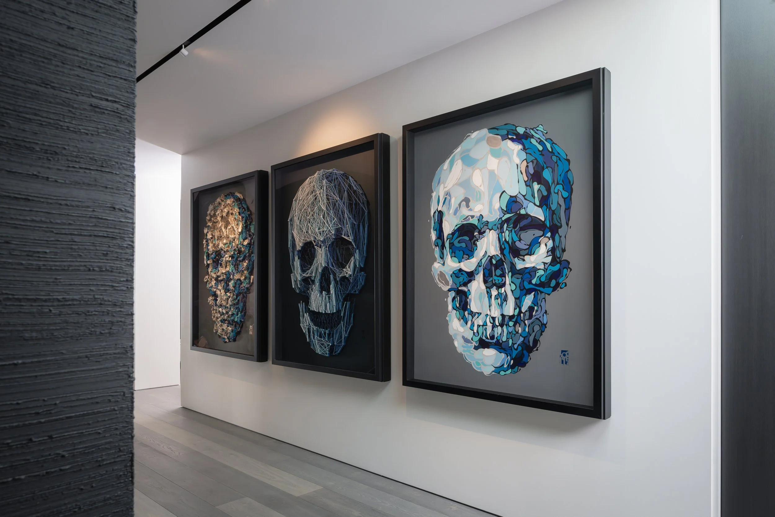 Three framed artworks of skulls with various styles hang on a white wall in an art gallery. The left skull appears abstract with textured materials, the middle skull looks like a geometric wireframe, and the right skull is stylized with blue and whit