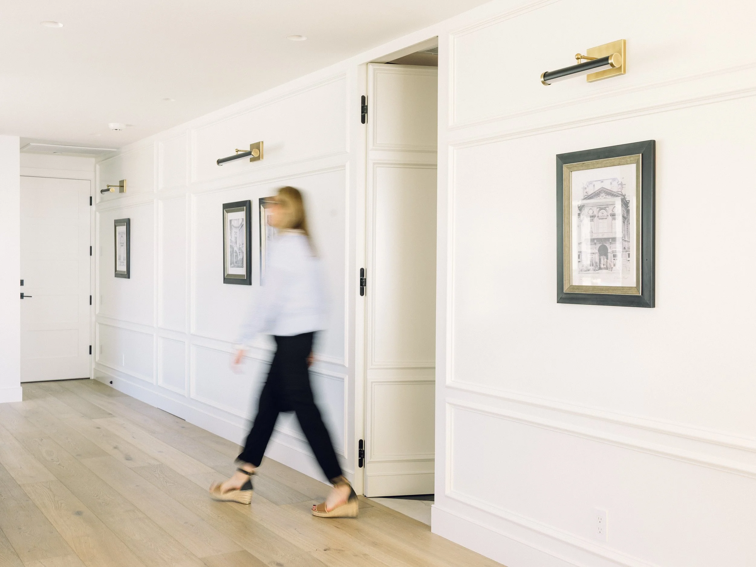 A woman walking through a hallway with white walls, wooden flooring, framed artwork, and modern wall-mounted lights.