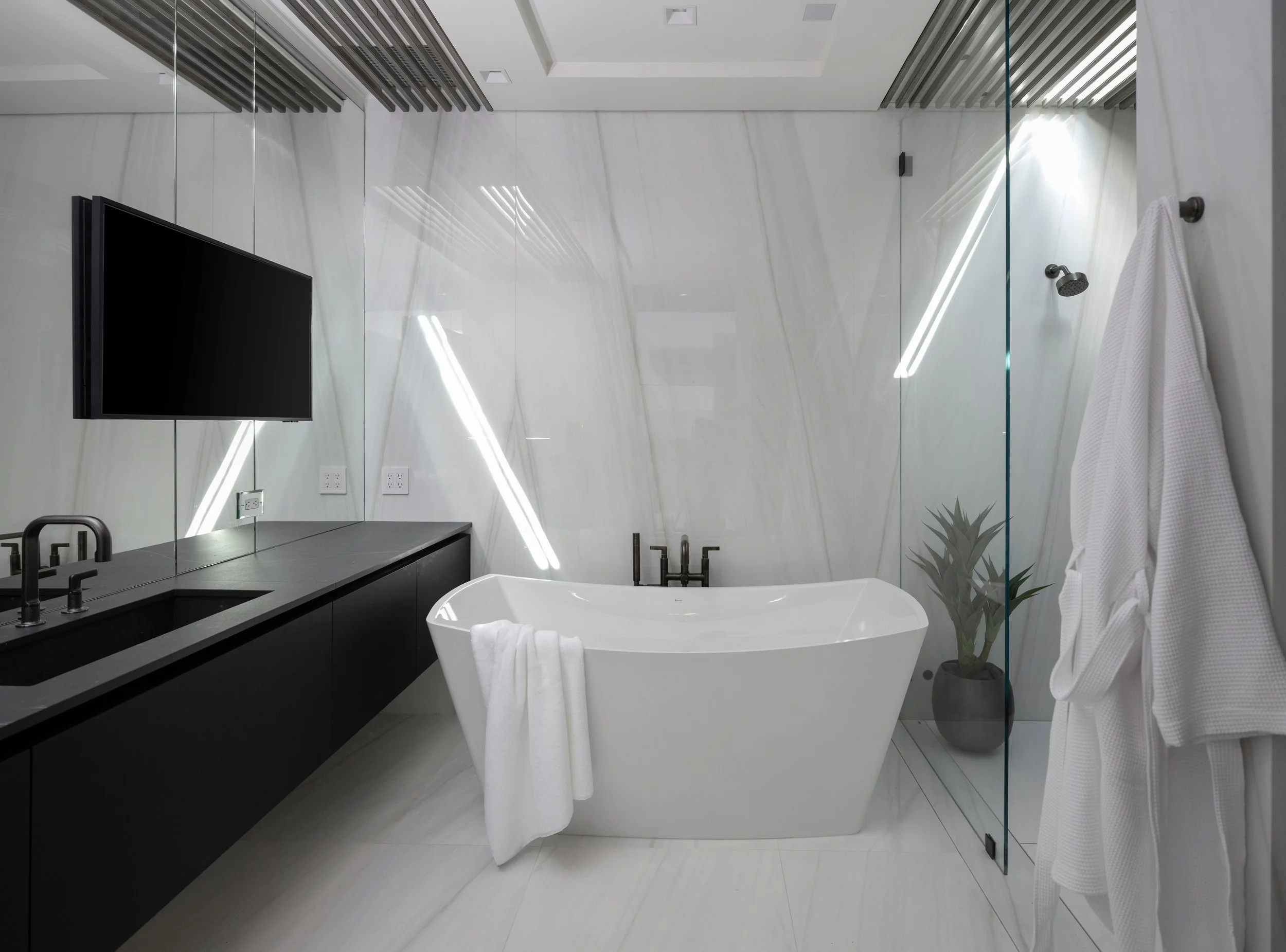 Modern bathroom with white bathtub, black fixtures, wall-mounted TV, potted plant, and white robes on rack.