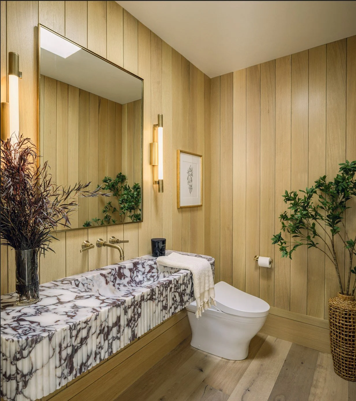 Modern bathroom with wood-paneled walls, marble sink, large mirror, framed artwork, potted plants, and gold fixtures.
