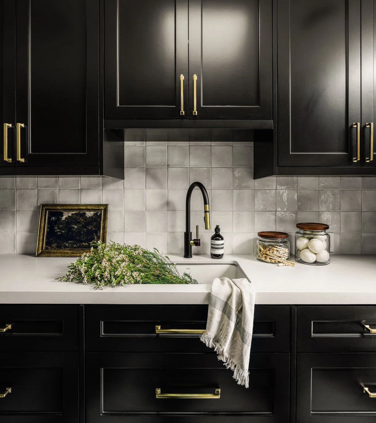 Black kitchen cabinets with gold handles, a white countertop, a black faucet, a sink with a bouquet of flowers, a framed picture, and jars with cotton balls and clothespins.