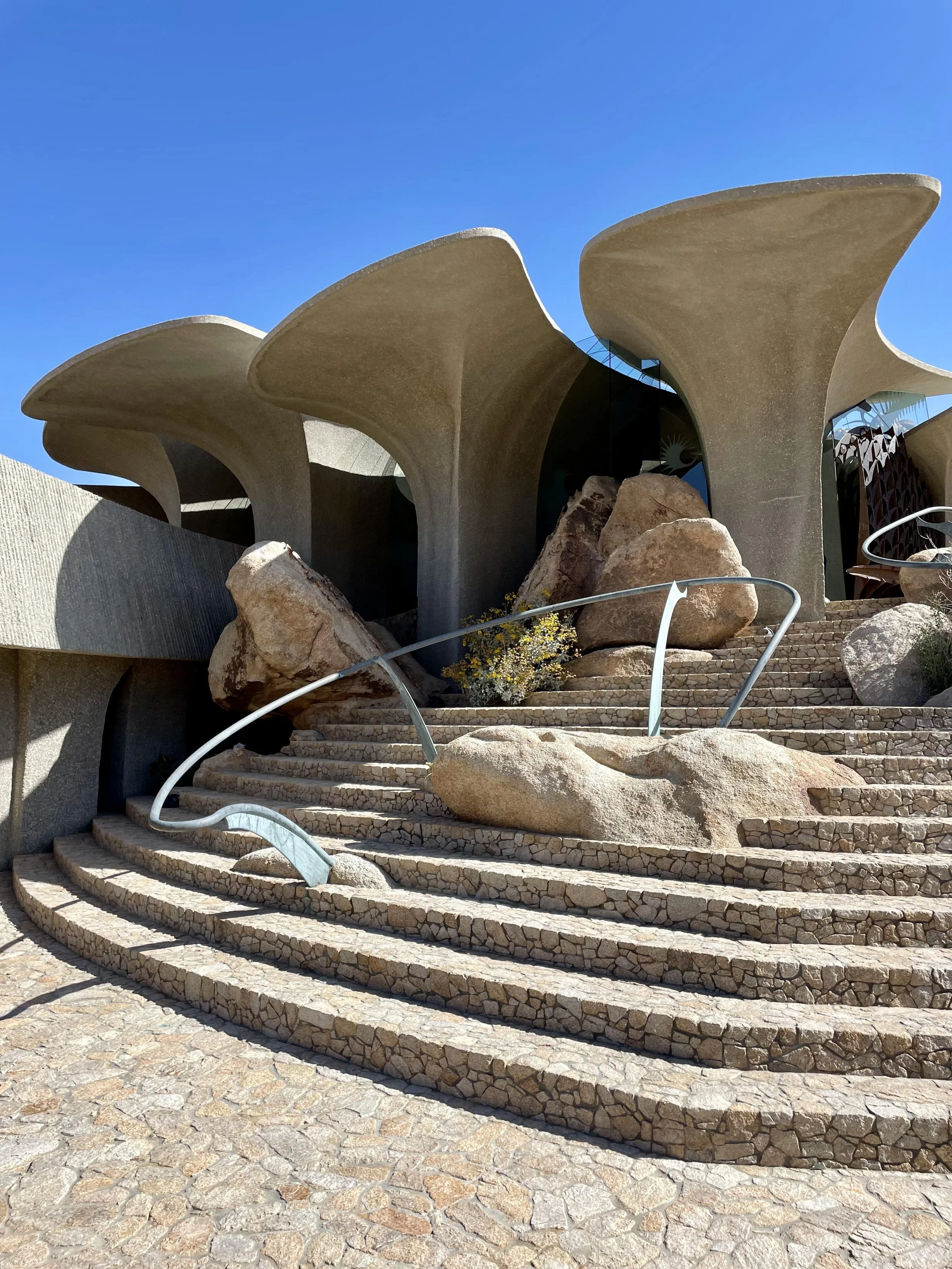 Unique modern building with large concrete flower petal-shaped roof structures, rocky landscape, and stone steps leading up to the entrance on a clear, sunny day.